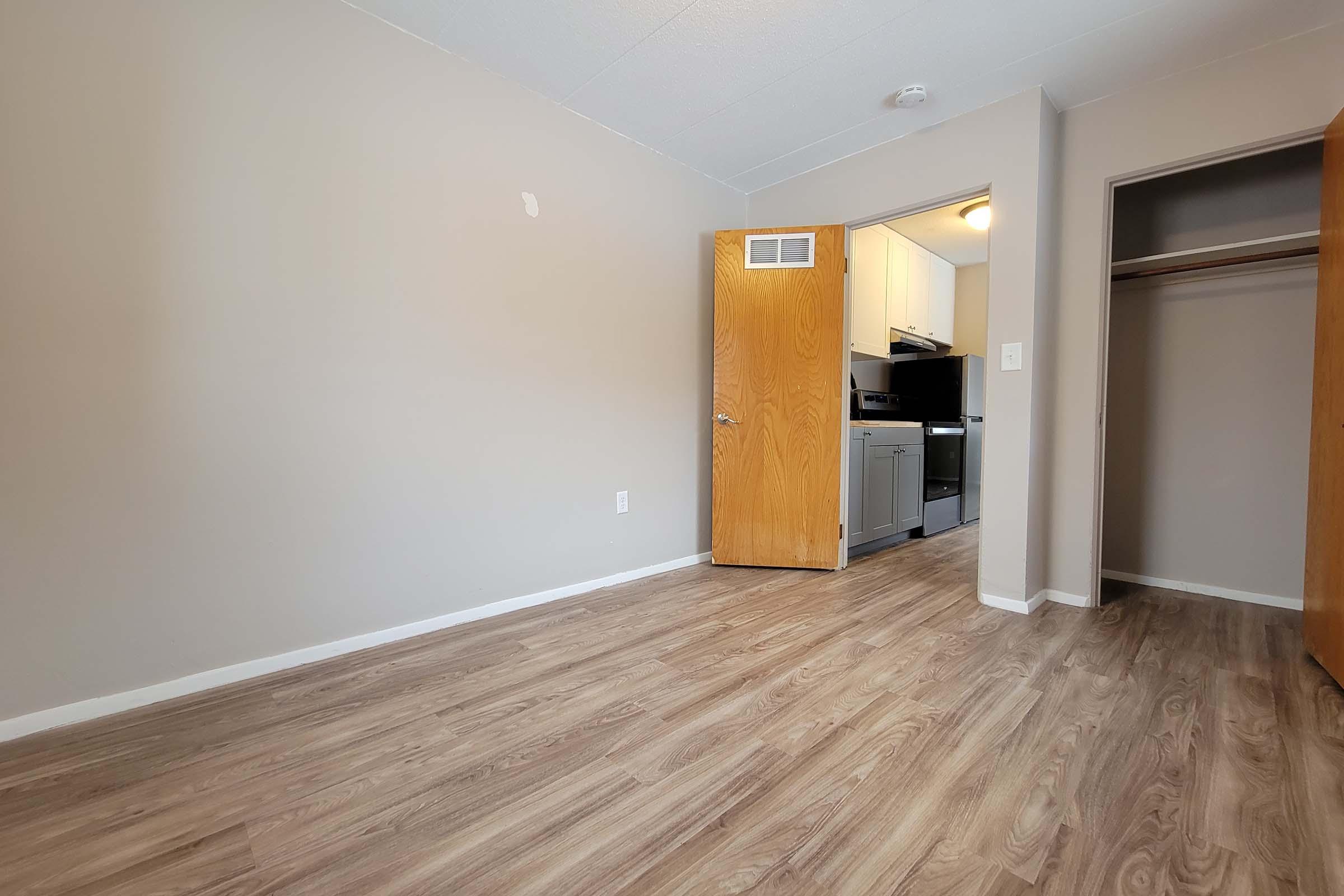 A small, empty room with light gray walls and wood laminate flooring. A doorway leads into a kitchen area visible in the background, featuring a stove and cabinets. There is a closet on the right with wooden doors partially open. Natural light is coming from the kitchen, illuminating the space.