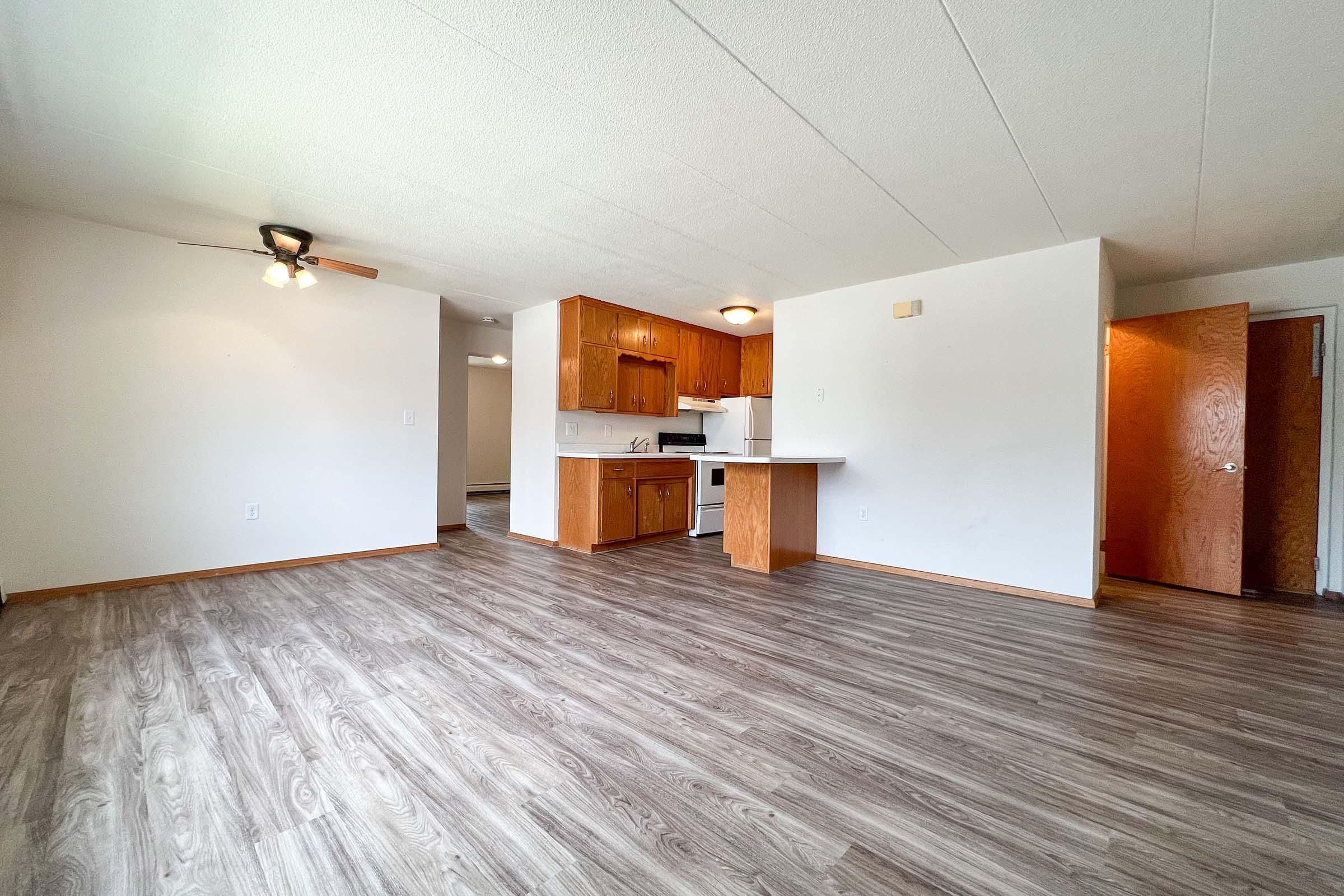 A spacious, empty living area with laminate flooring, featuring a ceiling fan and open access to a kitchen with wooden cabinets. There is a door leading to another room and a window letting in natural light. The overall atmosphere is bright and inviting, ideal for a cozy home.