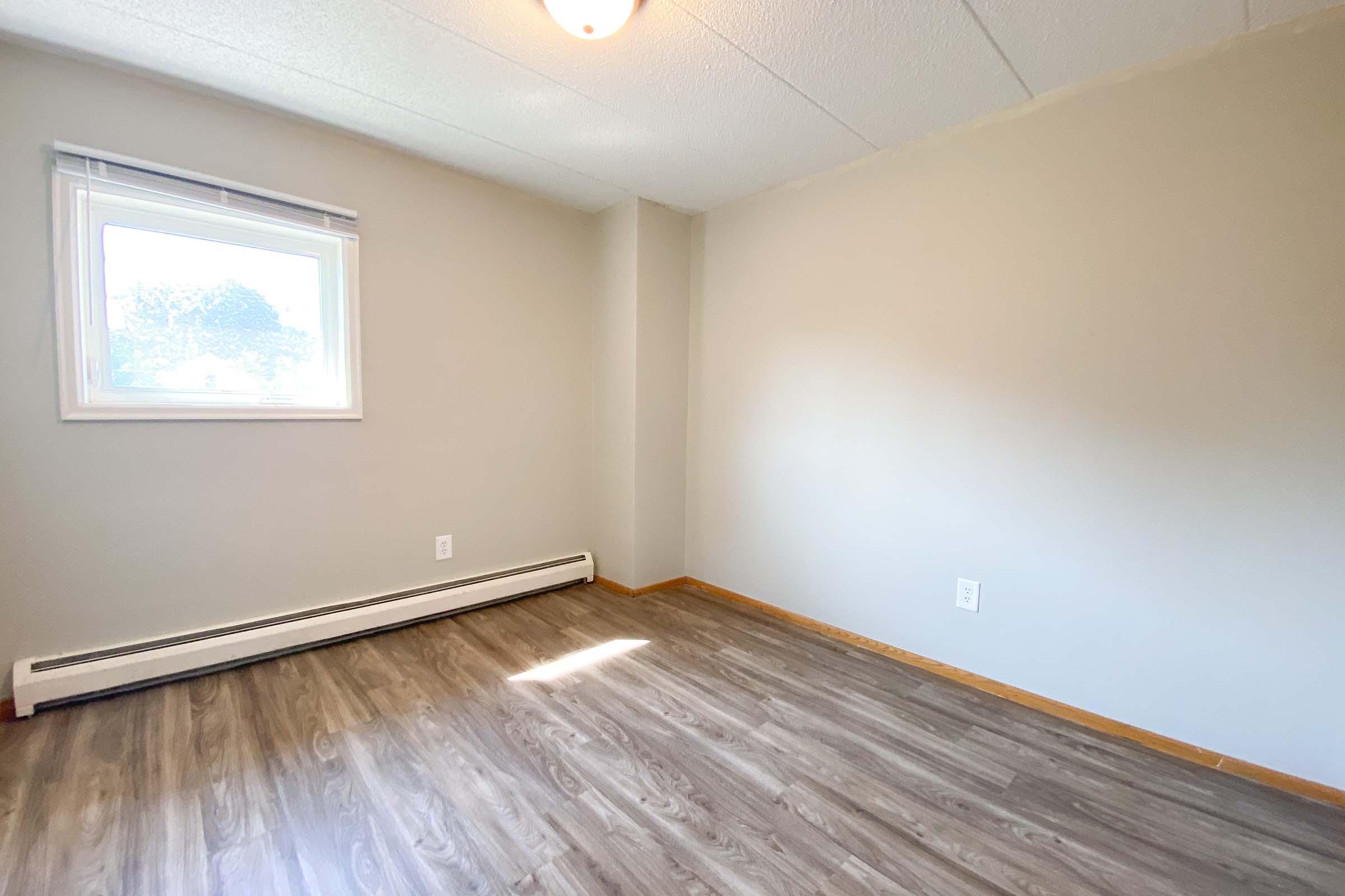 Empty room with light gray walls and a window allowing natural light. The floor features light brown laminate, while a white baseboard runs along the wall. A light fixture is visible on the ceiling. The room appears clean and spacious, suitable for various purposes.