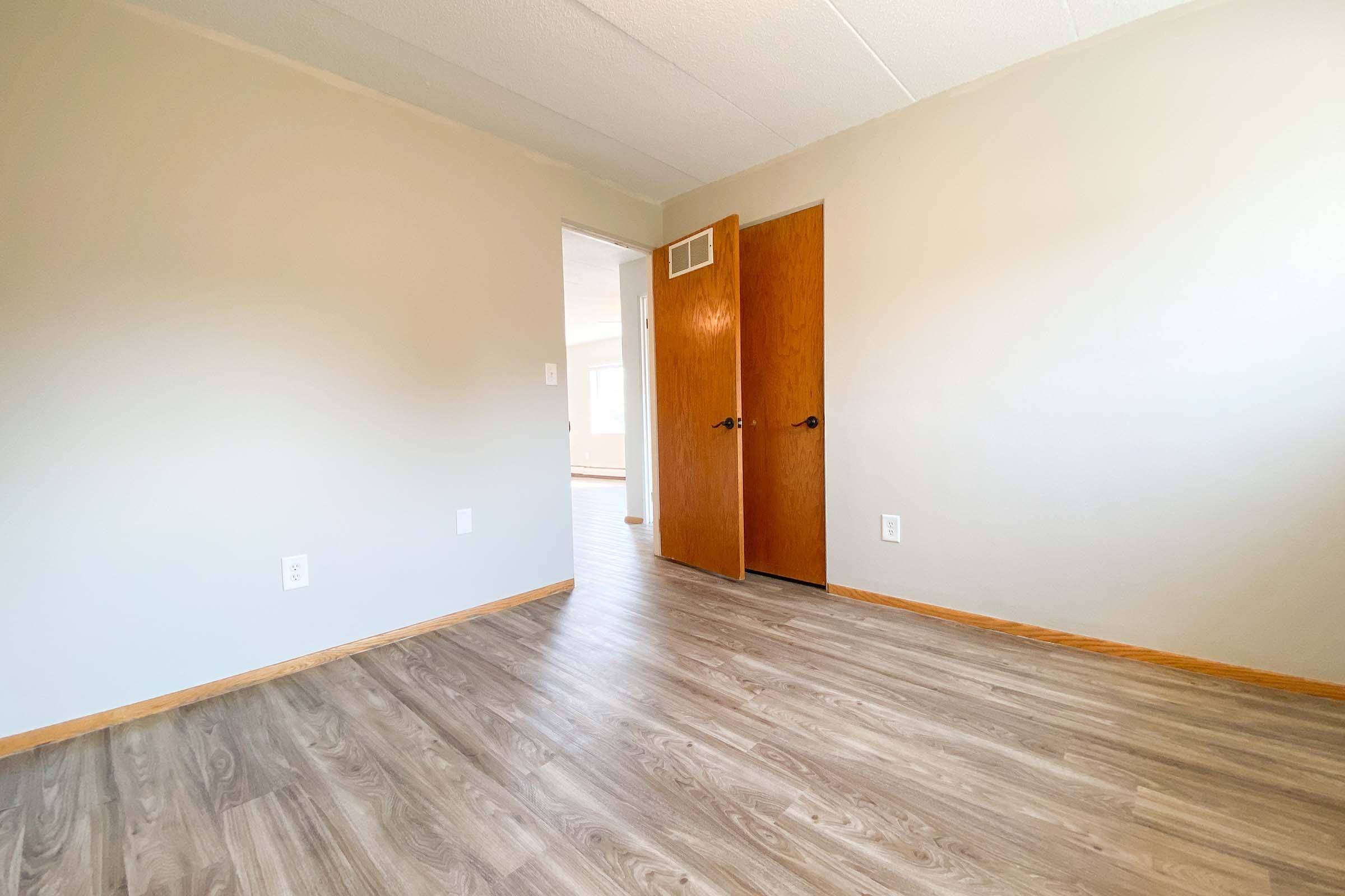 A vacant room with light gray walls, featuring a hardwood floor and a closed wooden door on the right. A doorway leads to another room, and there is natural light from a window in the background, creating a bright and spacious atmosphere.