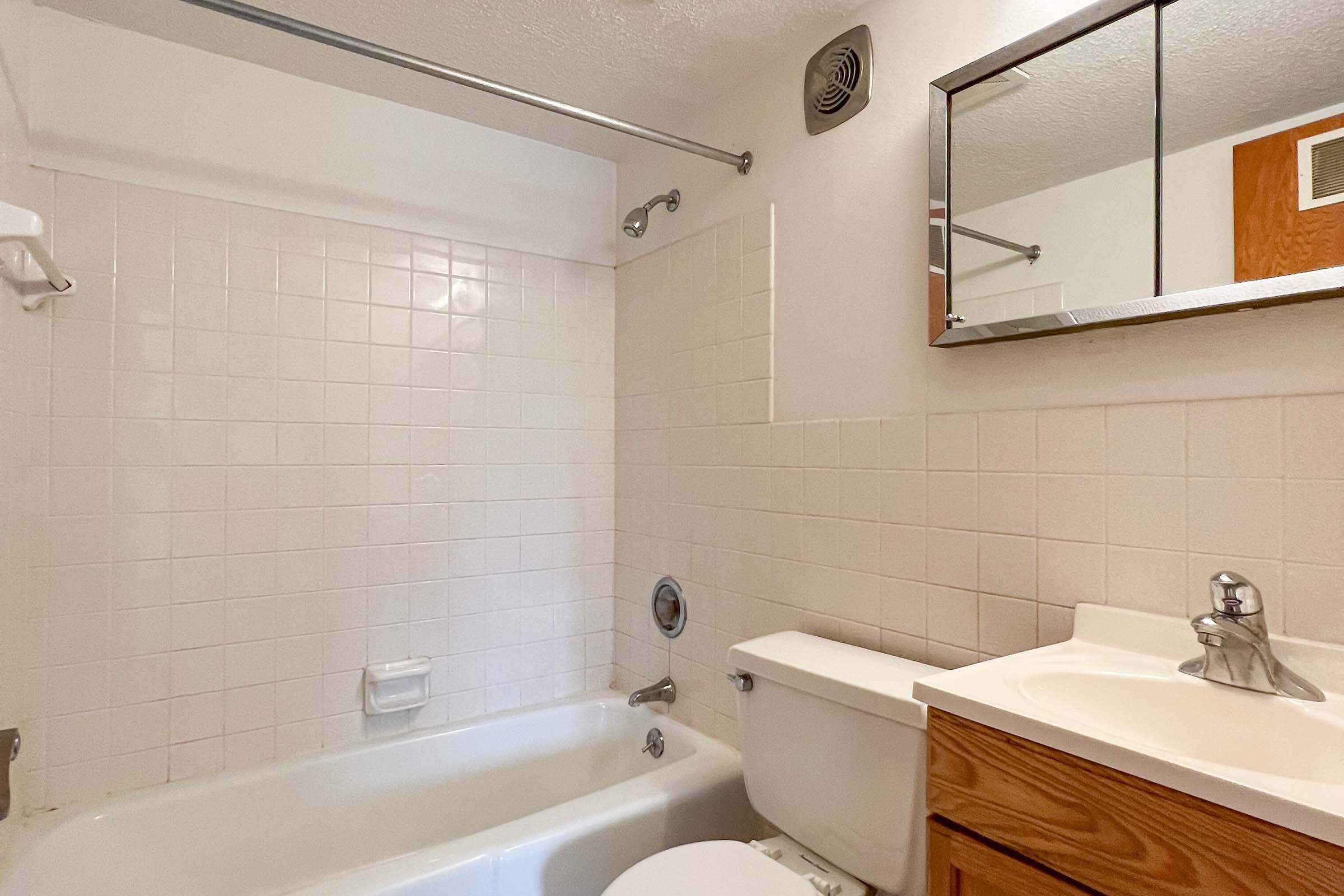 A clean and simple bathroom featuring a white bathtub, a small glass shelf, a mirror above the sink, and a toilet. The walls are covered with white tiles, and there is a wooden cabinet under the sink. The space is well-lit with a light fixture on the ceiling.