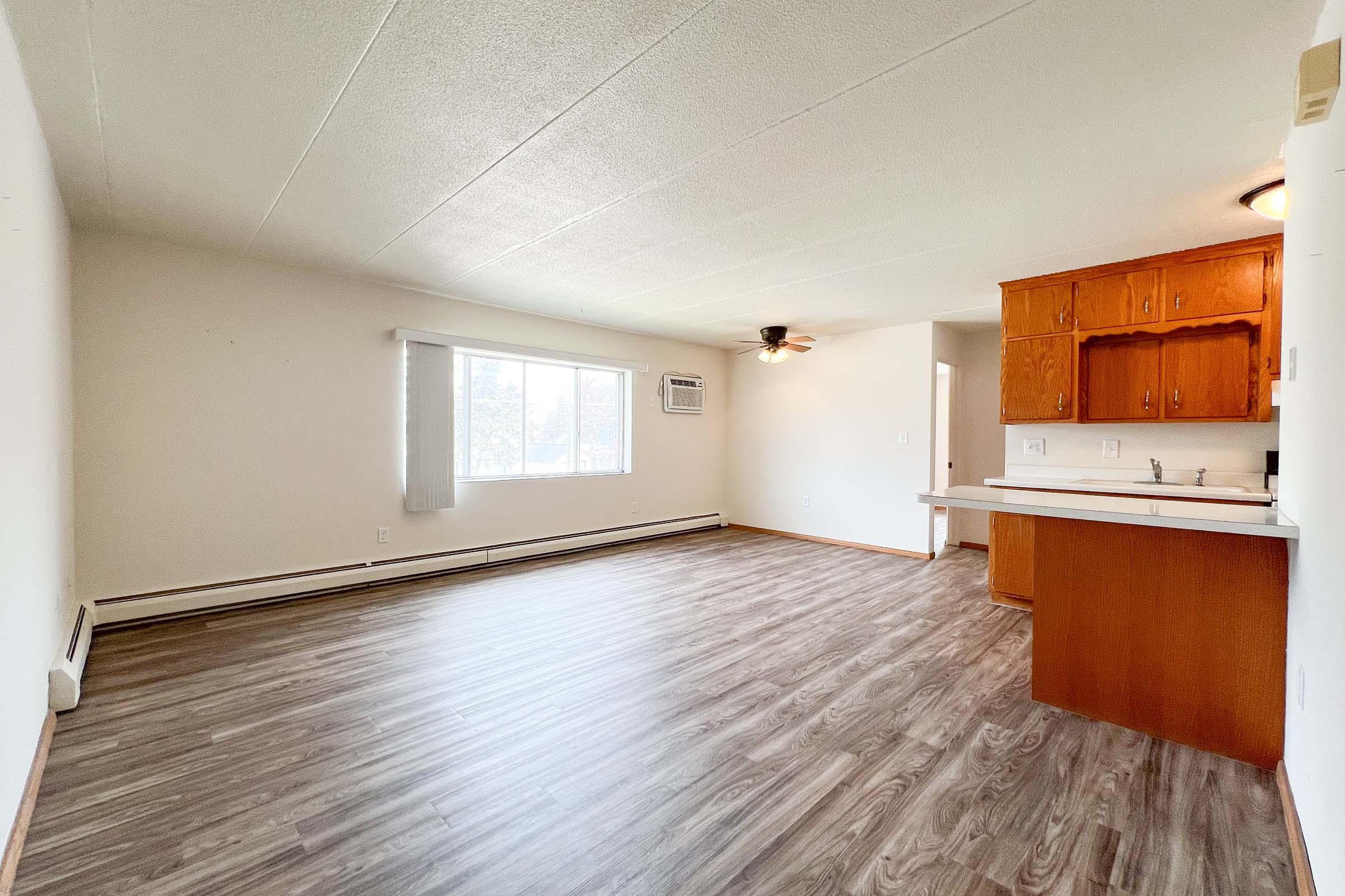 A spacious, empty room with light brown laminate flooring, a large window allowing natural light, an air conditioning unit, a ceiling fan, and wooden cabinetry in the corner. The walls are painted in a neutral color, creating a bright and open atmosphere.
