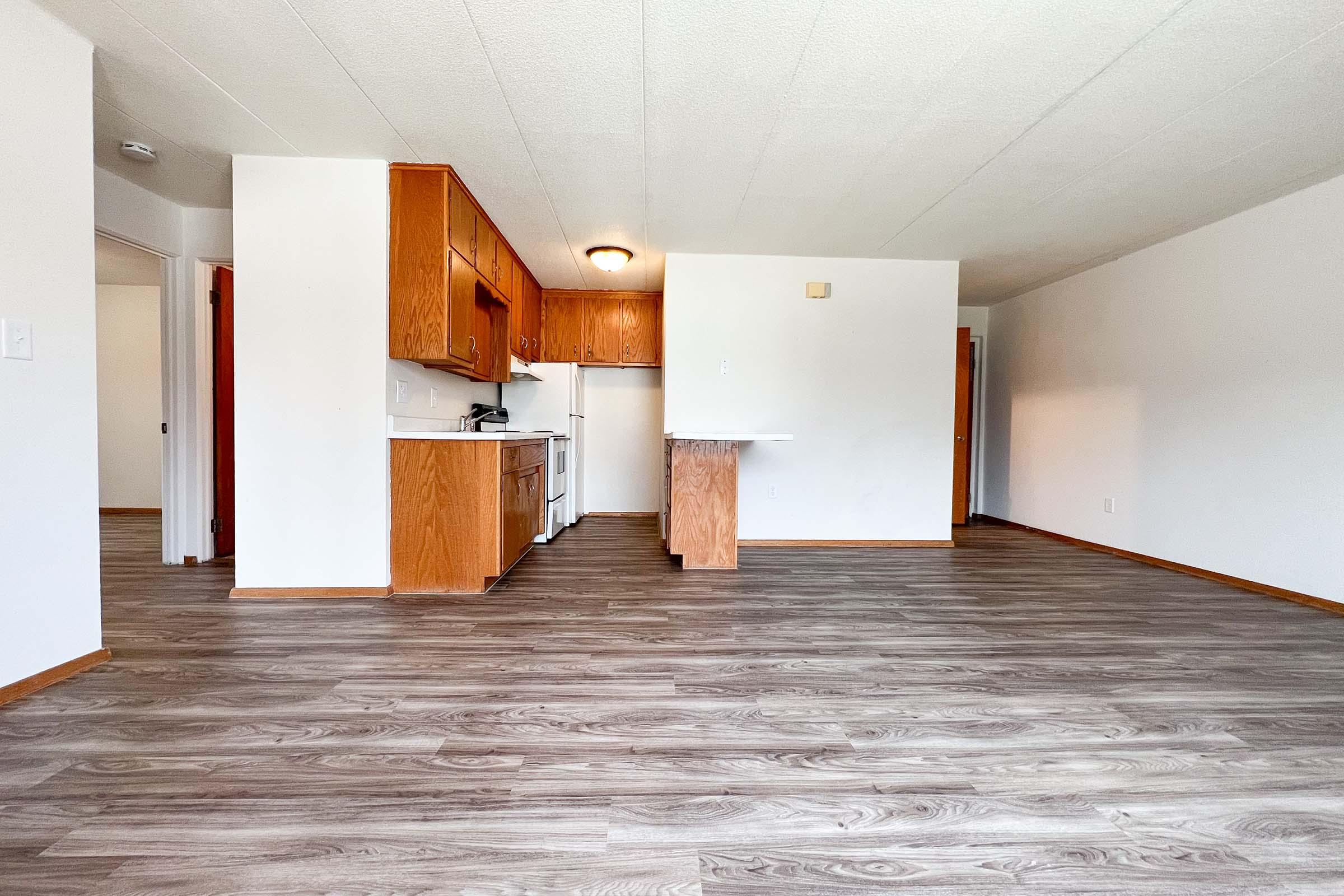 A spacious living area with light-colored walls and a wooden floor. The kitchen features wooden cabinets, a stove, and a refrigerator. A small countertop separates the kitchen from the living space. The room is well-lit, with a doorway leading to another room in the background on the left.