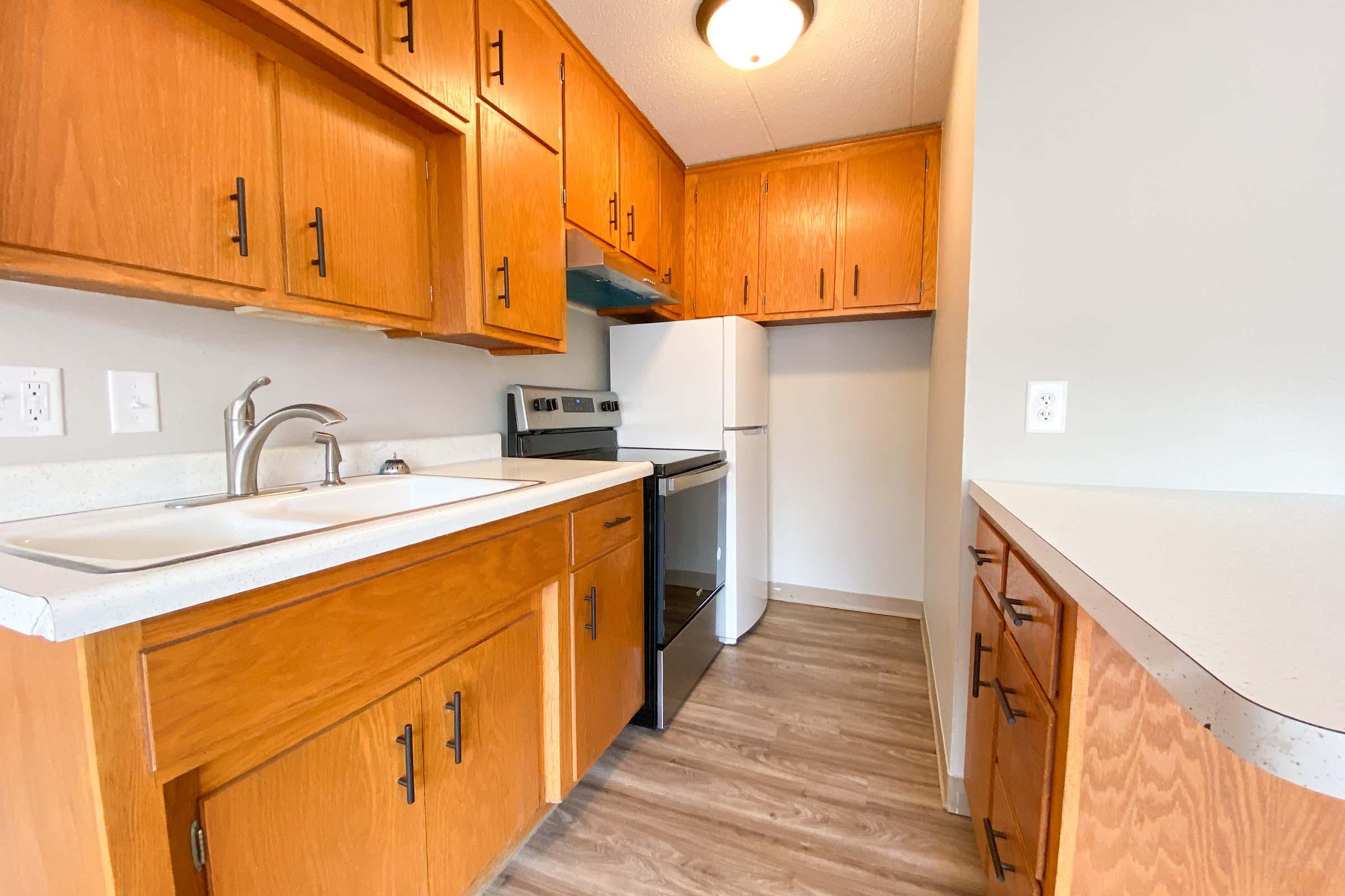 A modern kitchen featuring wooden cabinetry, a white countertop, a sink, and a stove. There is a black oven and a white refrigerator, with beige walls and laminate flooring. The area is bright, with overhead lighting, providing a functional and warm cooking space.