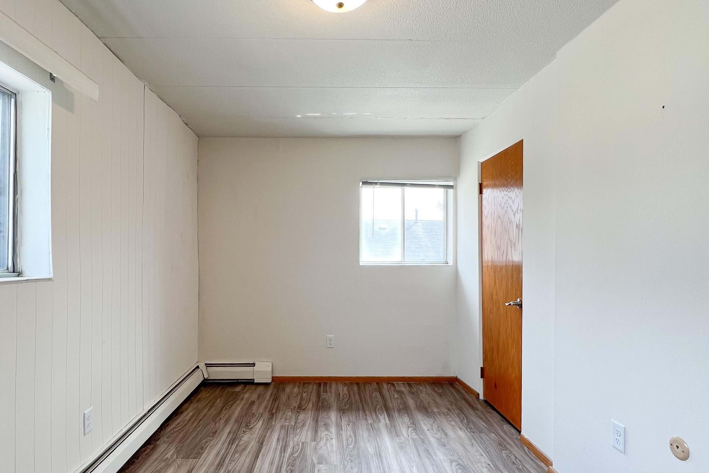A small, empty room with light-colored walls and a wooden door. It features a single window allowing natural light, an air vent on the floor, and laminate flooring. The ceiling is plain and white, and there are no furnishings or decorations visible in the space.