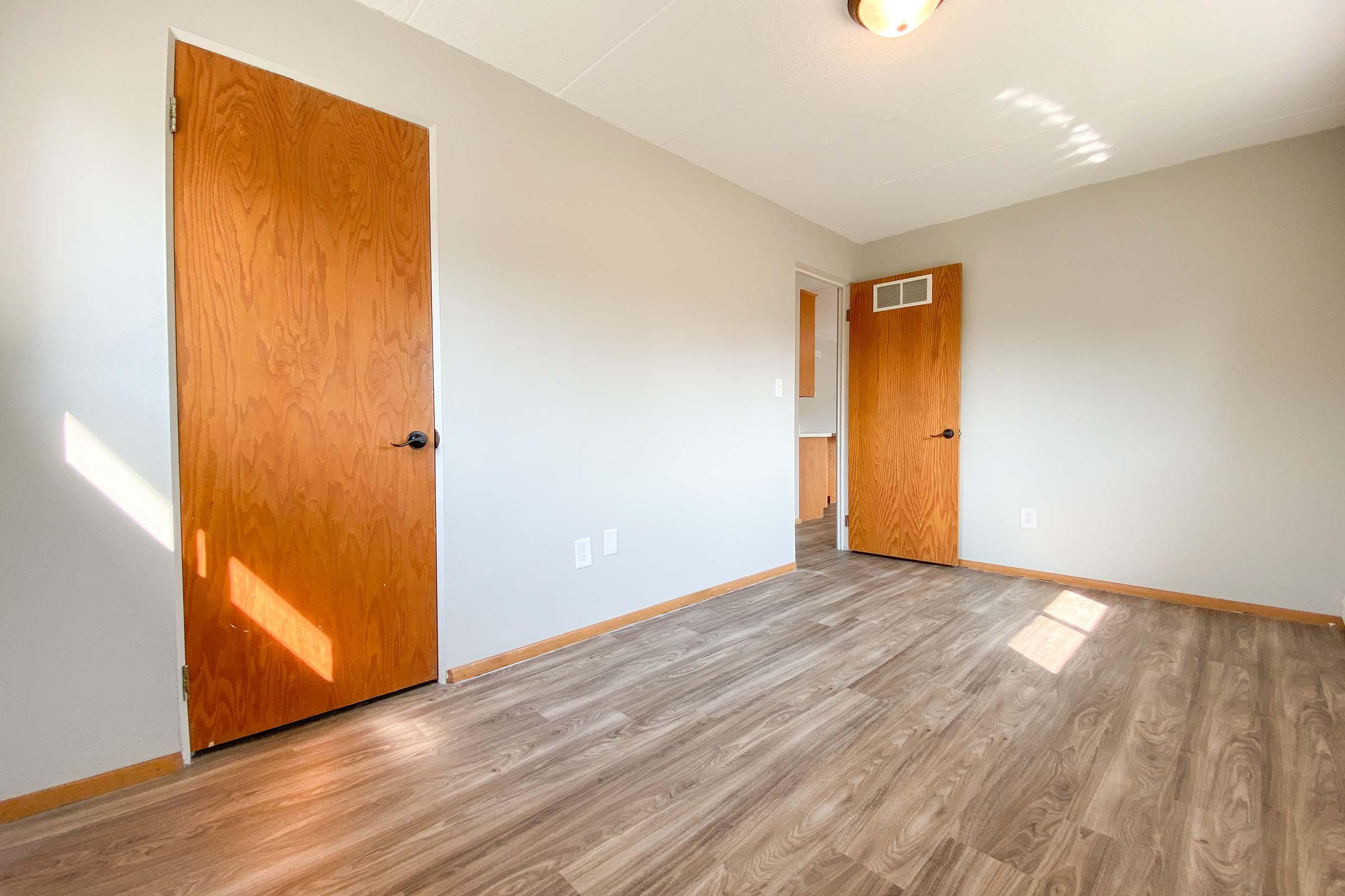 A spacious, empty room with light gray walls and a warm wood floor. Two wooden doors are visible—one on the left and one on the right. Natural light enters the room, creating a bright atmosphere. There is a small hallway visible through the door on the right.