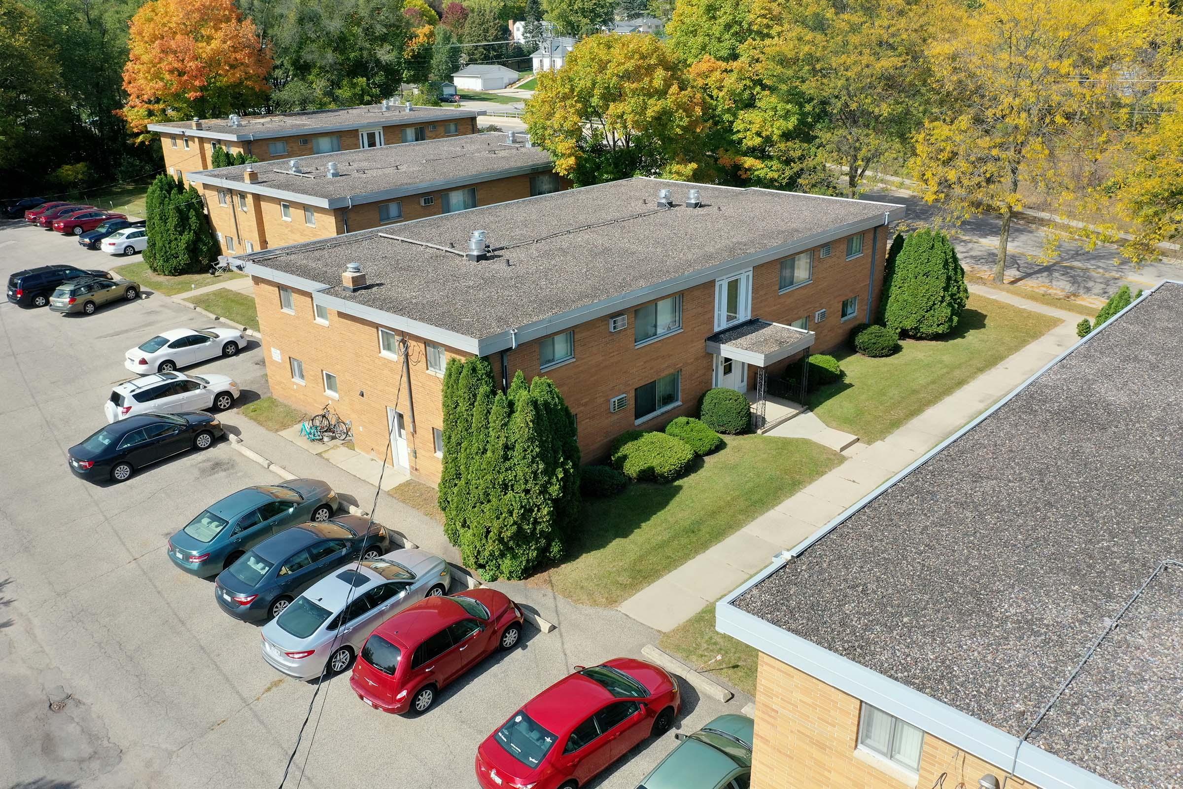 Aerial view of a residential apartment complex with multiple brick buildings, surrounded by green lawns and landscaped shrubs. Several parked cars occupy the lot, showcasing a mix of colors. Trees with autumn foliage are visible in the background, indicating a suburban setting.
