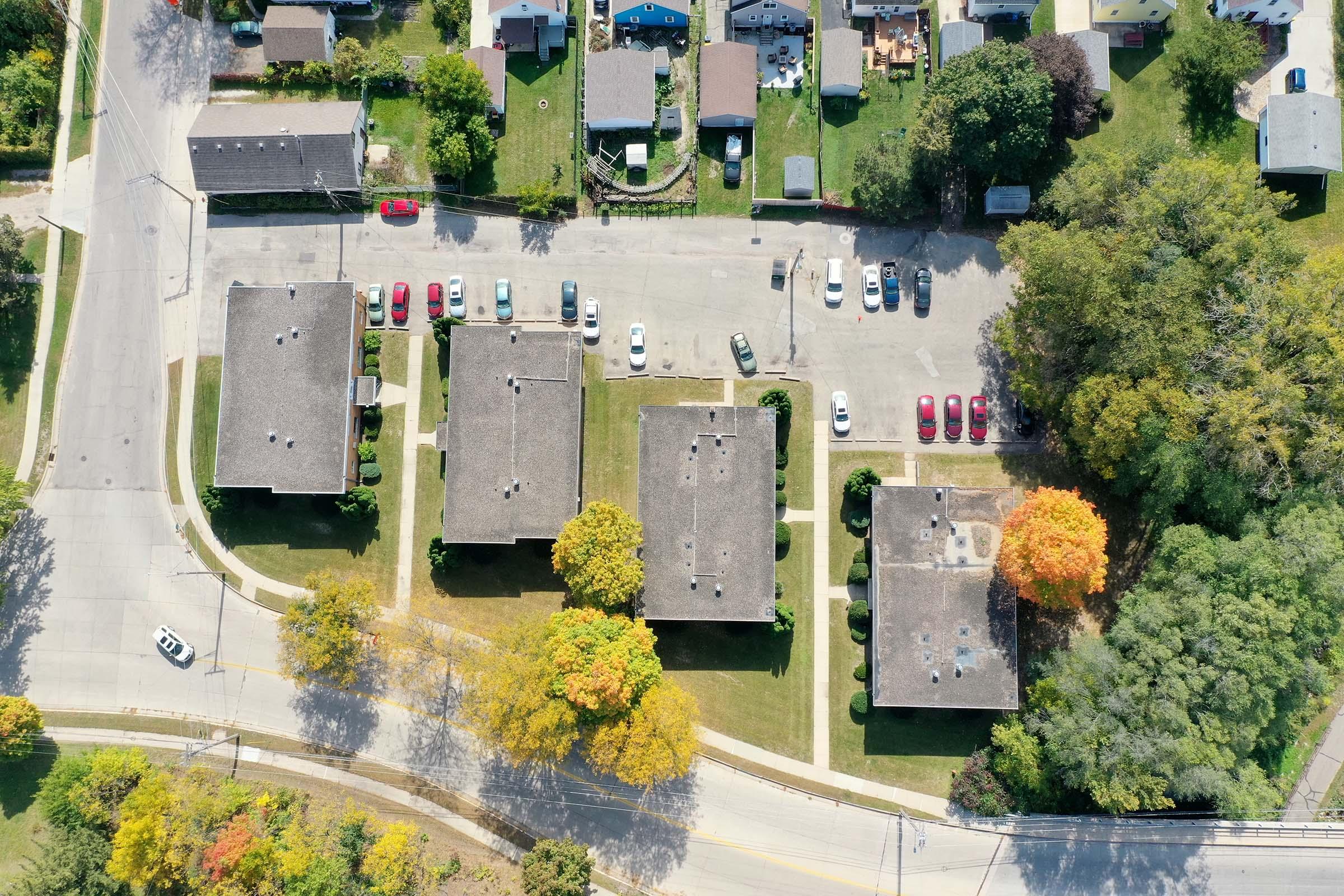 Aerial view of a residential area featuring several apartment buildings arranged in rows, surrounded by green lawns. The parking lot is visible with numerous vehicles parked, and trees with autumn foliage in the foreground. Streets and sidewalks intersect the area, showing a blend of urban and natural elements.