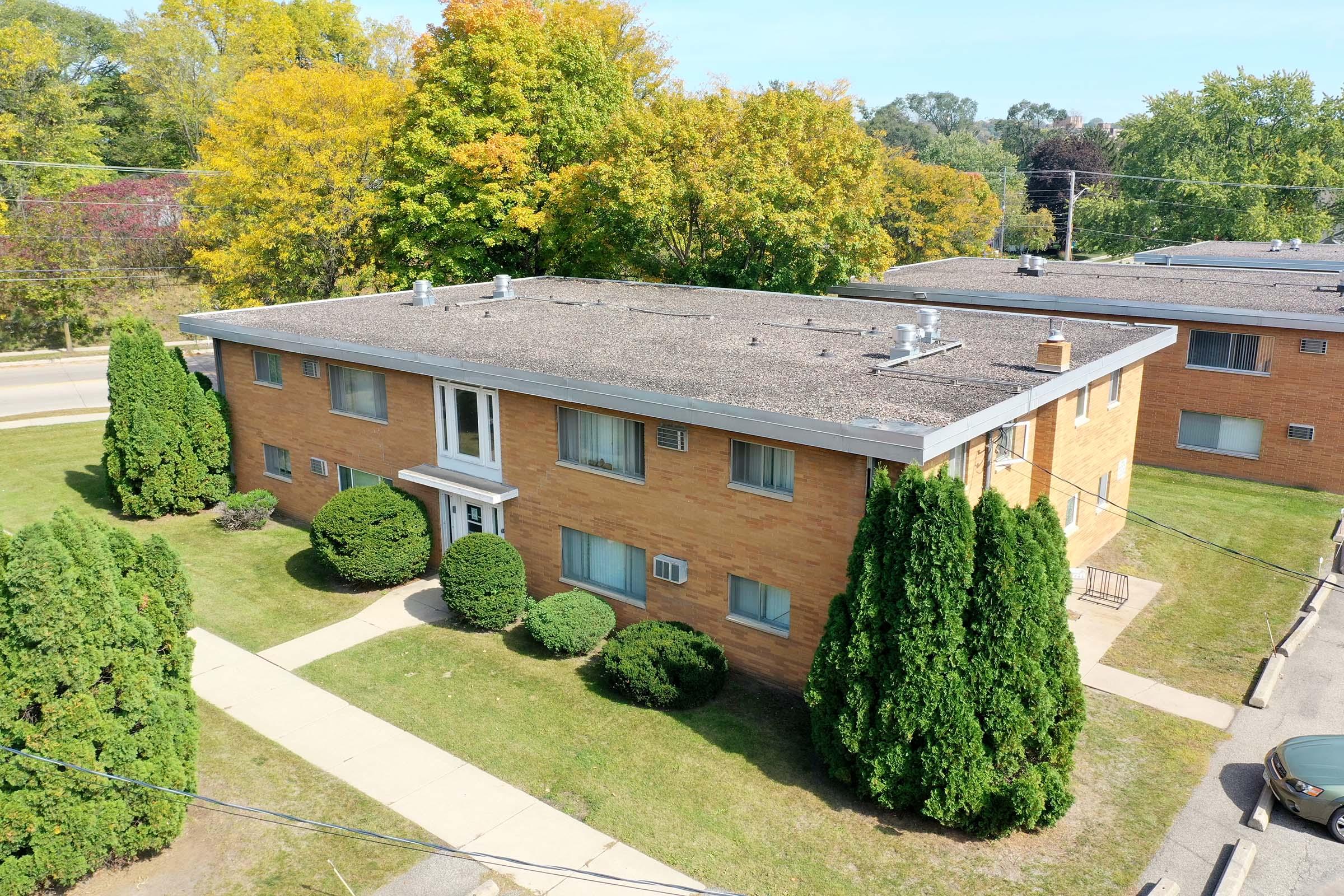 Aerial view of a two-story brick apartment building surrounded by well-maintained greenery. The building features several windows and air conditioning units, with neatly trimmed shrubs in front and a paved walkway leading to the entrance. Trees with autumn foliage can be seen in the background.