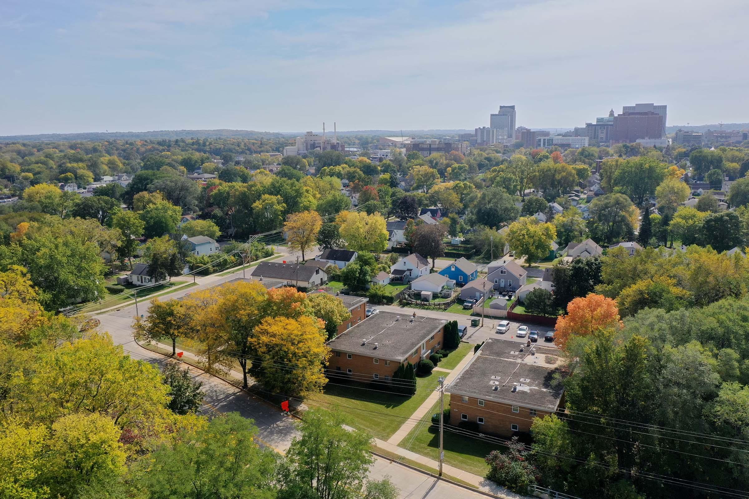 Aerial view of a suburban area featuring rows of houses with colorful autumn foliage. In the background, a city skyline with tall buildings is visible under a clear sky. The scene captures a blend of urban and natural landscapes, showcasing trees in vibrant fall colors.