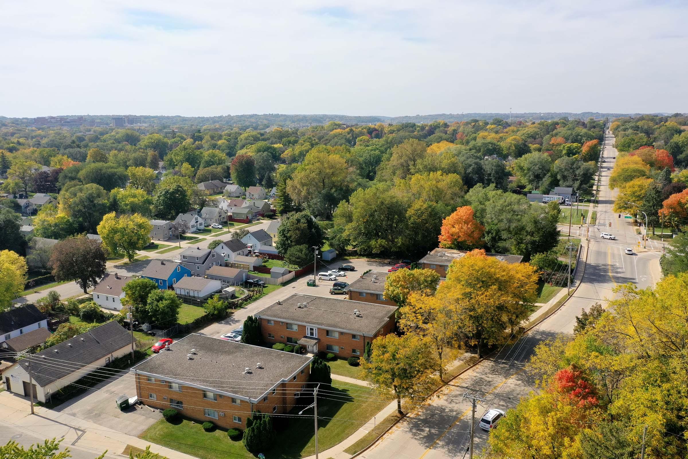 Aerial view of a suburban neighborhood in autumn, featuring a mix of residential buildings and tree-lined streets. The trees display vibrant fall colors, while houses with varied rooftops are visible. The scene captures the essence of a tranquil suburban setting surrounded by nature.