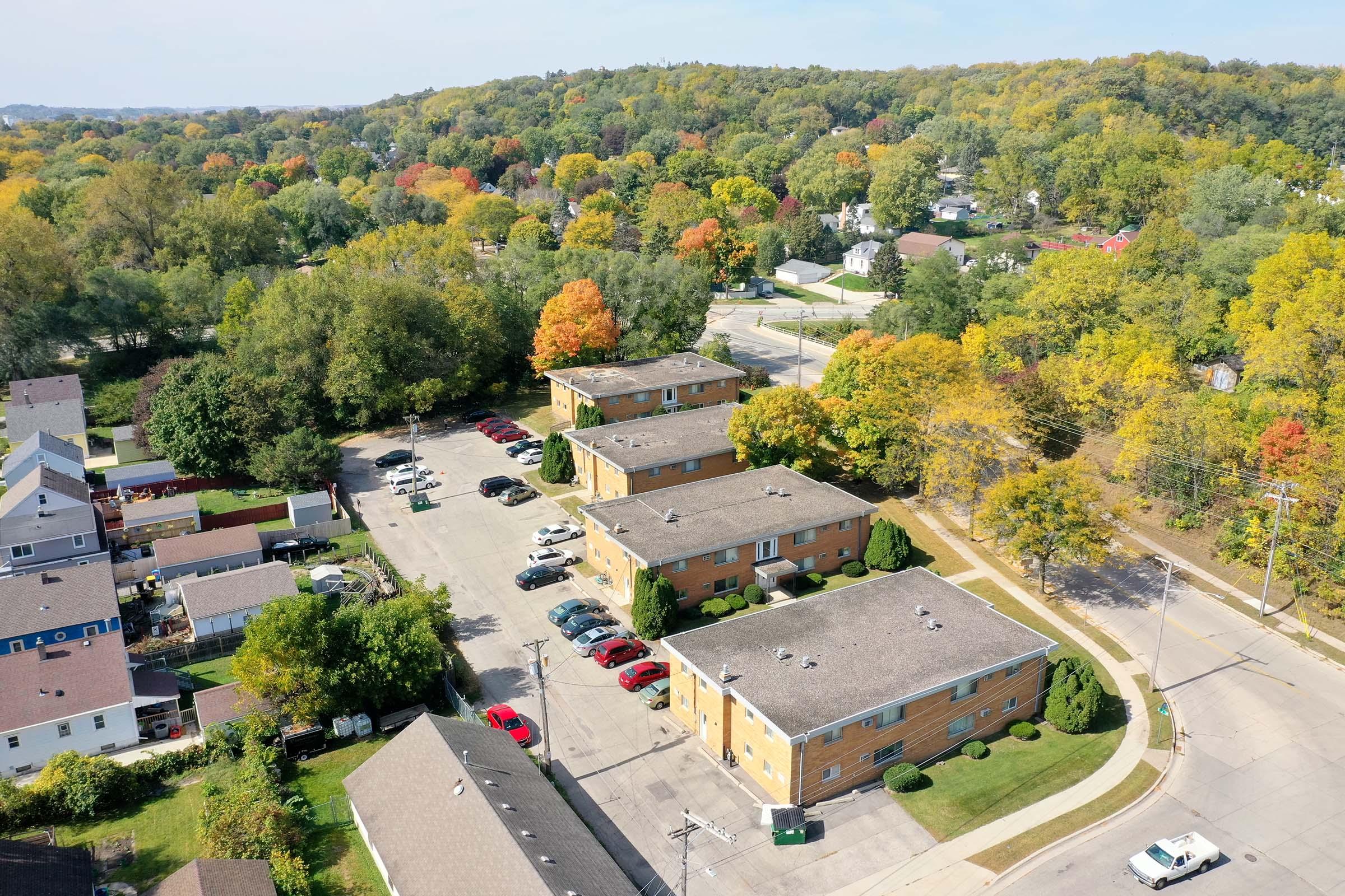 Aerial view of a residential area with several apartment buildings, surrounded by trees displaying autumn foliage. The scene includes parked cars in a lot, winding streets, and a mix of green and colorful trees, indicating a vibrant fall landscape.