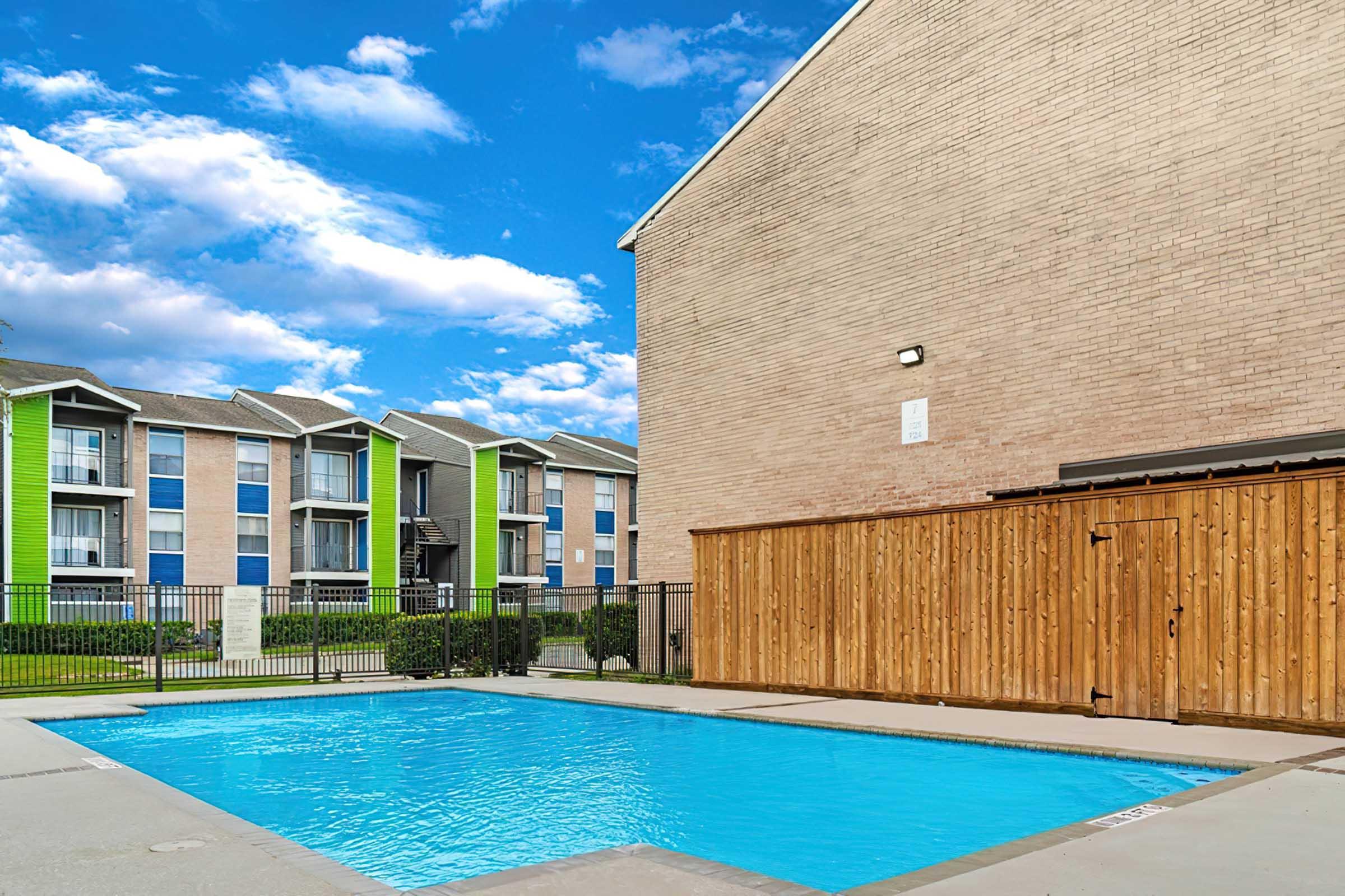 A clear blue swimming pool surrounded by a concrete deck, with a wooden fence nearby. In the background, there are multi-story apartment buildings featuring green and blue accents under a partly cloudy sky.
