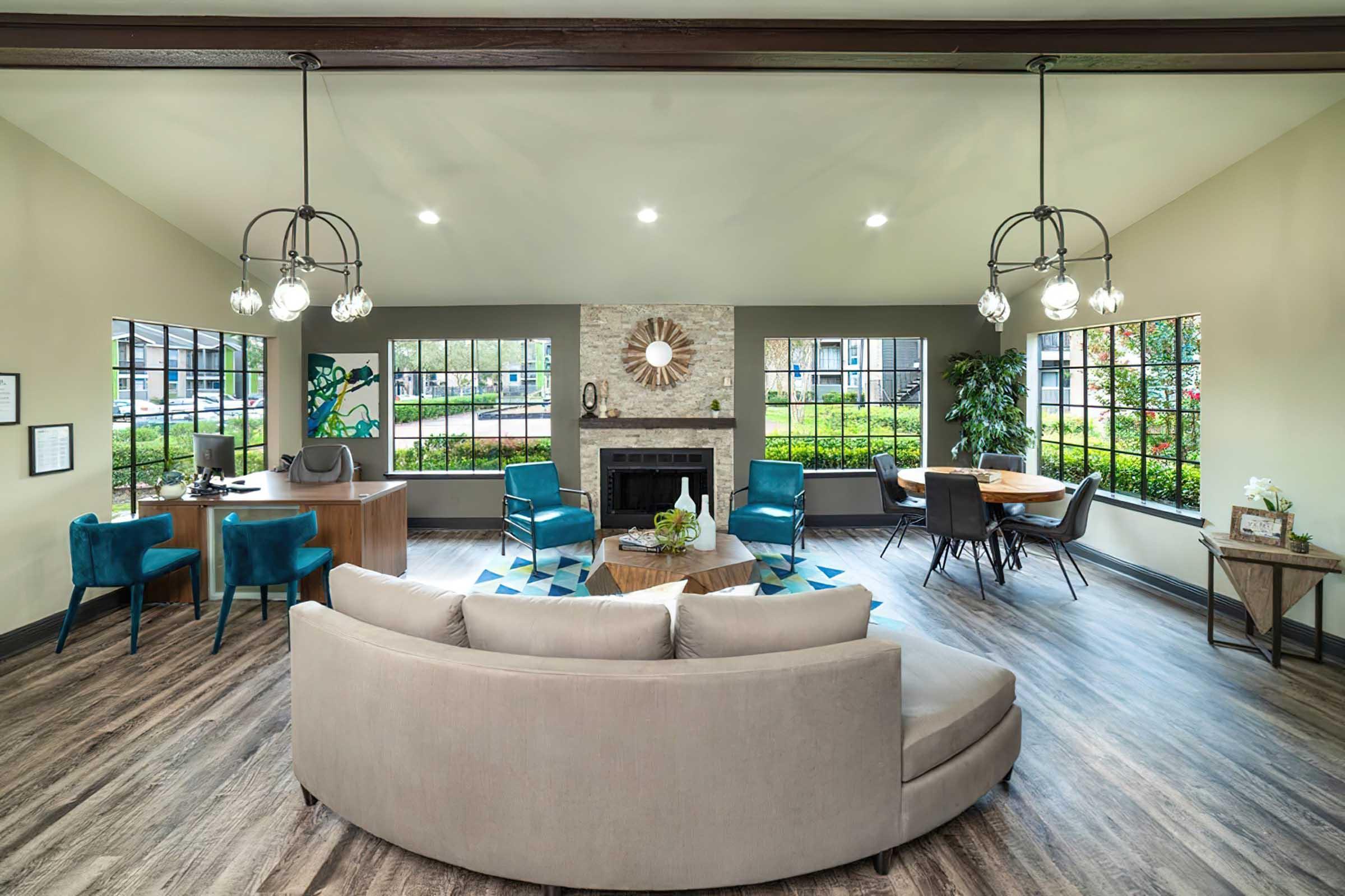 A spacious, modern living area featuring a circular sofa, two teal chairs, a wooden table, and a stone fireplace. Large windows provide natural light and a view of greenery outside. Decorative elements include a wall mirror and artwork on the walls. The flooring is a stylish wood finish.