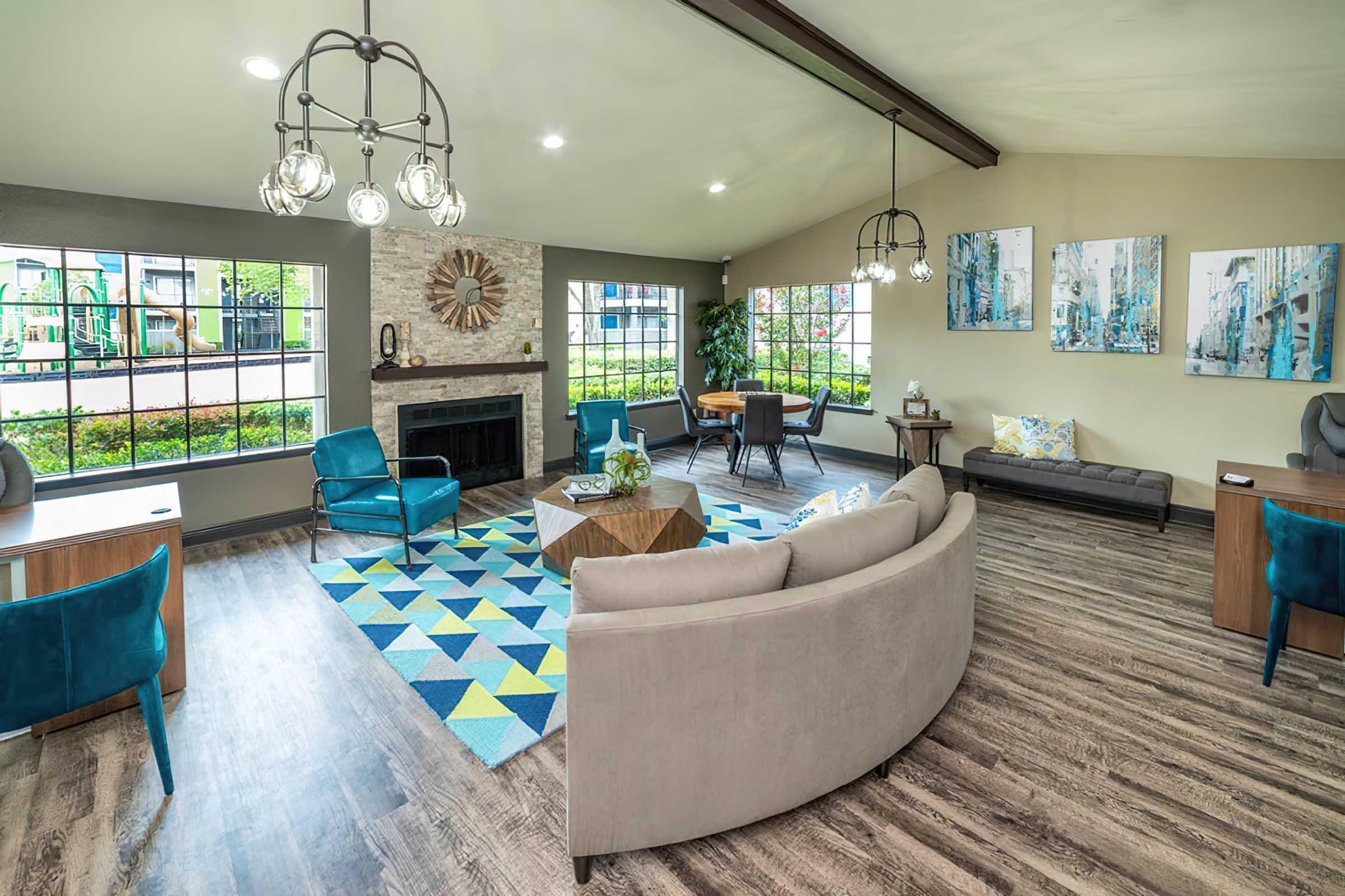 A cozy and modern living area featuring large windows, a fireplace, and stylish furniture. There are two blue chairs, a gray sofa, and wooden tables. The floor is covered with a colorful geometric area rug. Wall art and decorative plants add a touch of elegance to the space. Natural light brightens the room.