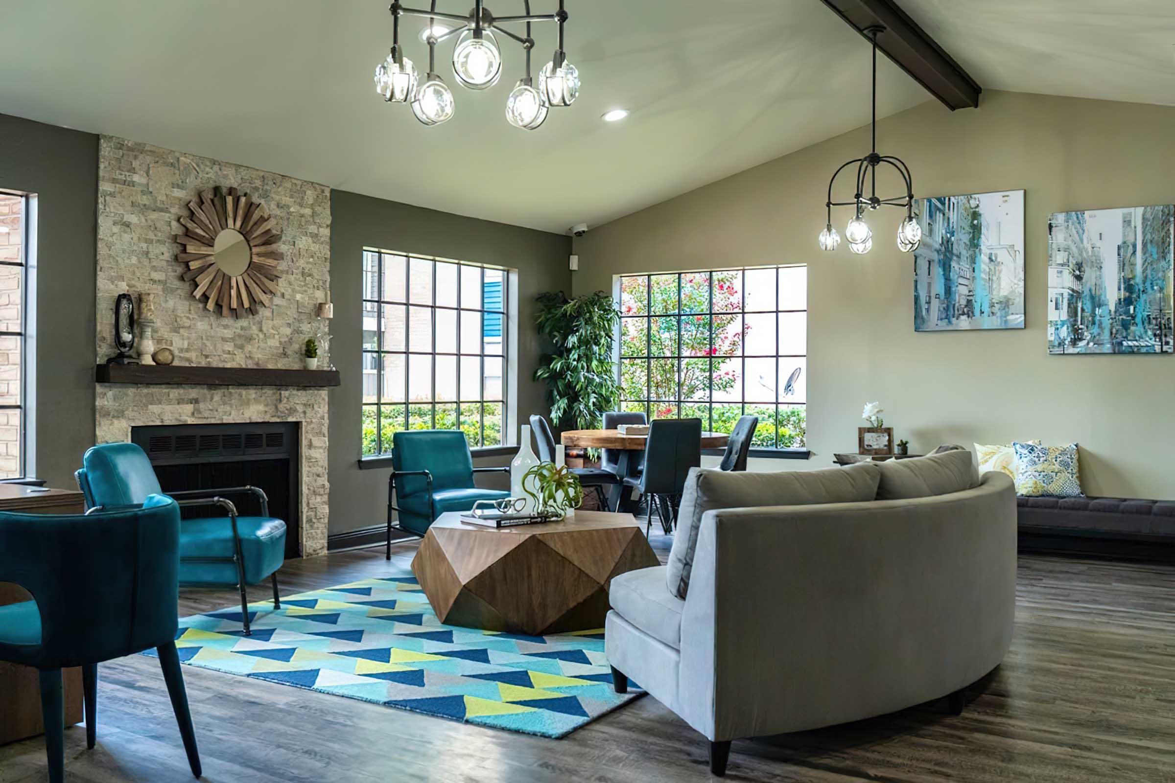 A modern living room featuring a stone fireplace, large windows, and contemporary furnishings. The space includes blue and gray seating, a geometric area rug, a wooden coffee table, and decorative artwork on the walls, creating a bright and inviting atmosphere.