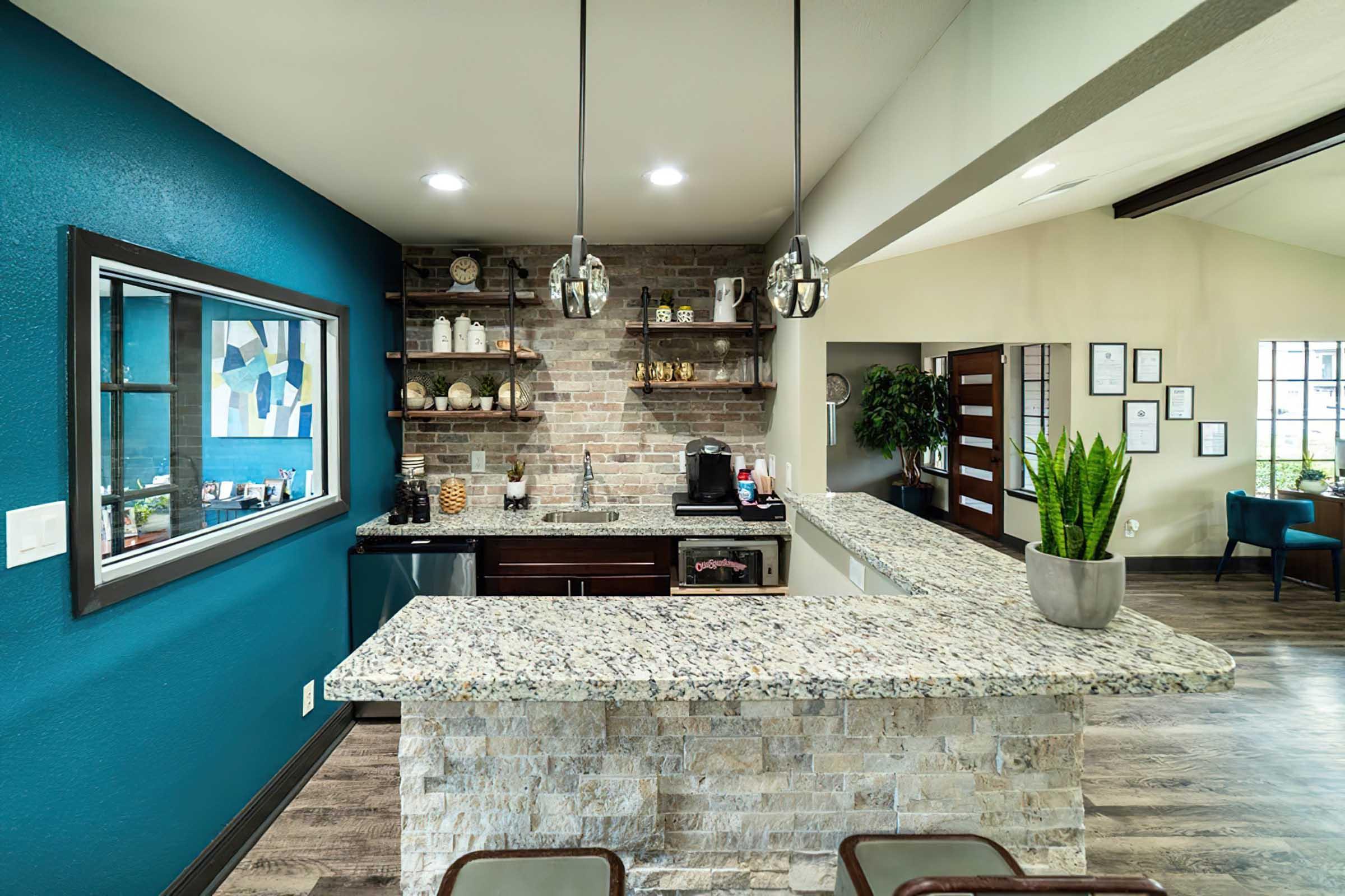 Modern kitchen with a granite countertop and stone accent wall. Features open shelving with decorative items, a coffee station, and pendant lighting. The walls are painted in vibrant blue and neutral tones, and there are plants for decor. Part of the space includes large windows and a cozy seating area.