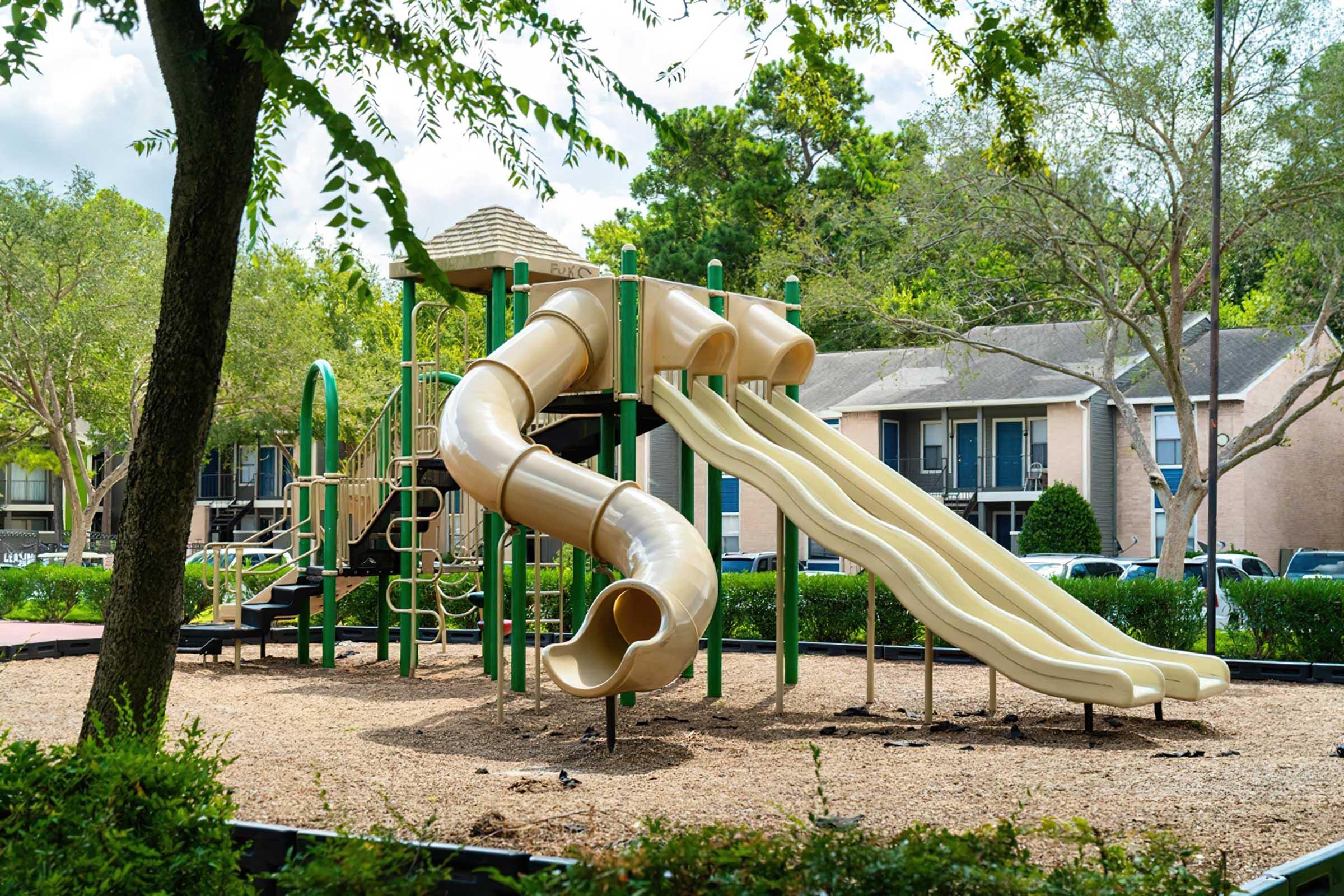 A playground featuring a large play structure with two slides—one curvy and one straight—surrounded by trees and well-maintained greenery. In the background, there are several residential buildings. The area is sunny, creating a cheerful and inviting atmosphere for children.