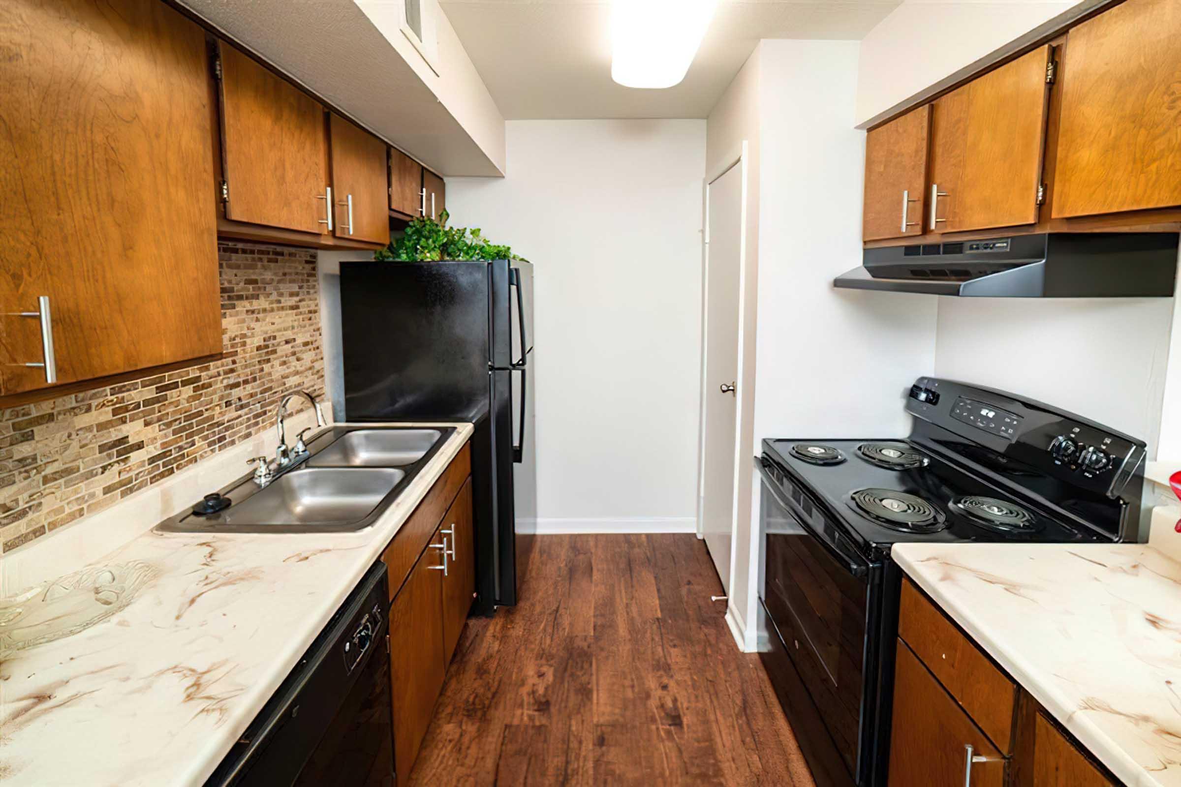 Modern kitchen featuring wooden cabinets, stainless steel sink with double basins, and black appliances including a refrigerator and stove. Countertops have a light marbled design, and there's a decorative backsplash. The floor is a warm wood tone, creating a cozy atmosphere.