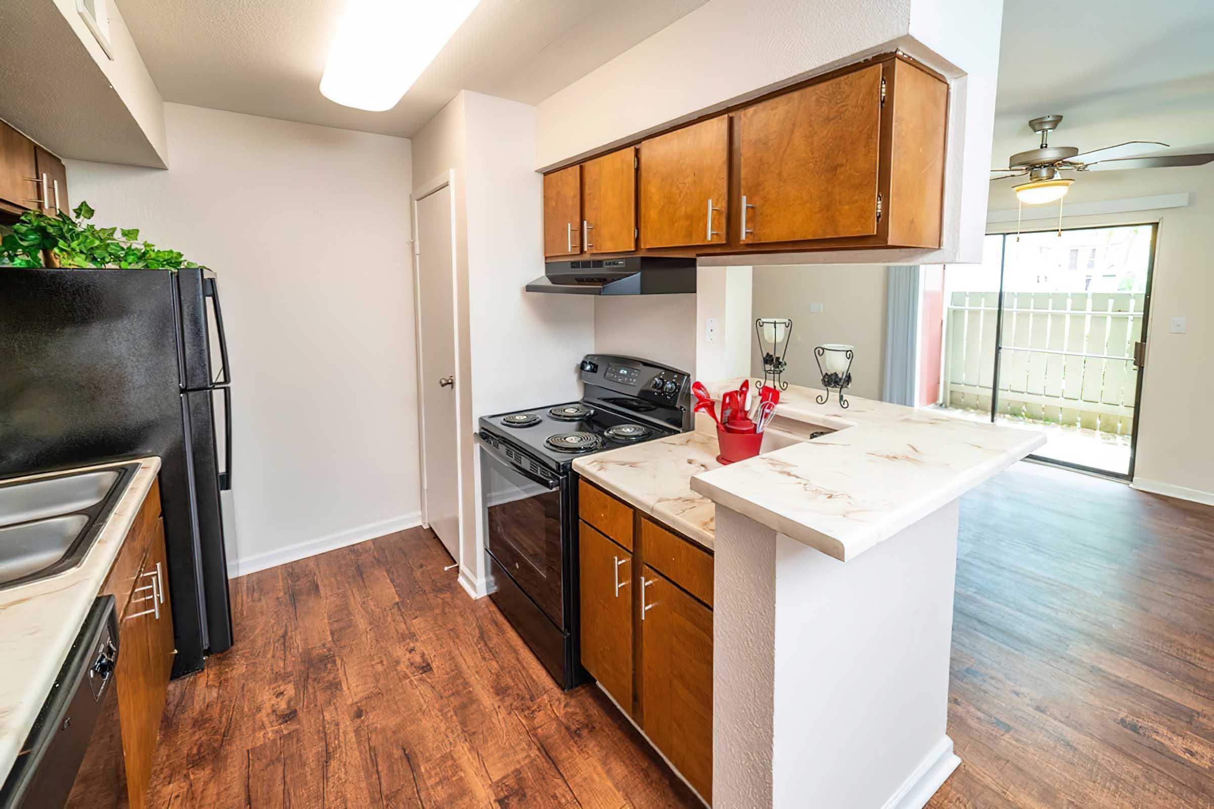 Modern kitchen featuring wooden cabinets, black appliances, and a countertop opening to a living area. The kitchen has a sink, stove, and ample space, with a door leading to an outdoor patio area in the background. The flooring is a warm wood finish, creating a cozy ambiance.