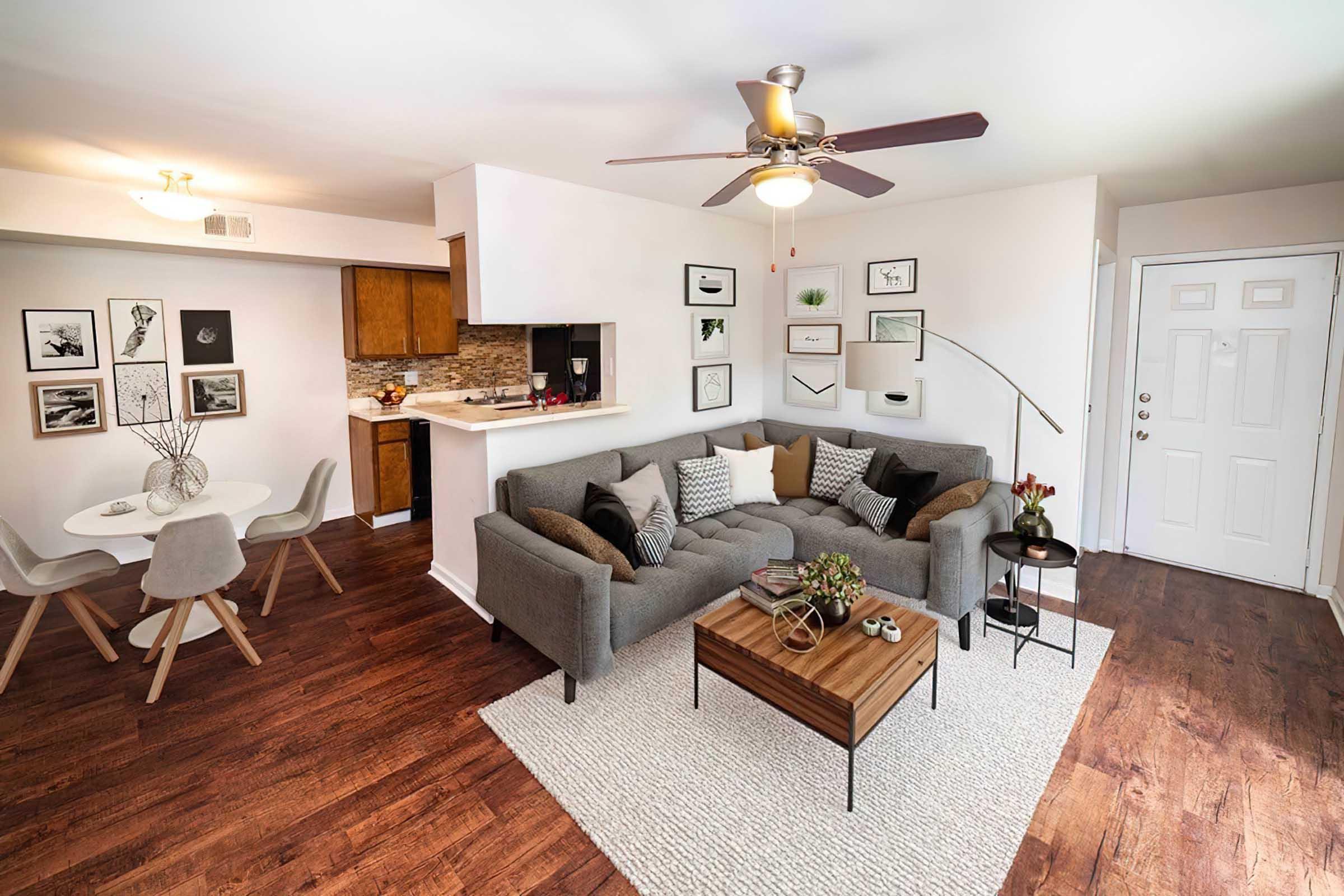 A modern living room featuring a gray sectional sofa with decorative pillows, a wooden coffee table, and a cozy area rug. There's a dining area with a round table and chairs in the background. The space includes framed artwork on the walls and a ceiling fan, with a doorway leading to another room.