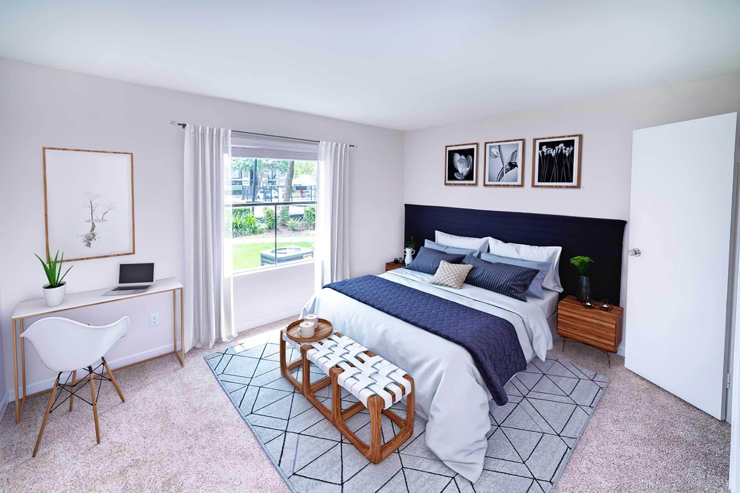 A modern bedroom featuring a bed with gray and navy bedding, a stylish bench at the foot, and a small desk. The walls are adorned with framed art, and a large window provides natural light, offering views of greenery outside. The decor is minimalistic and contemporary, with neutral tones and soft textures.