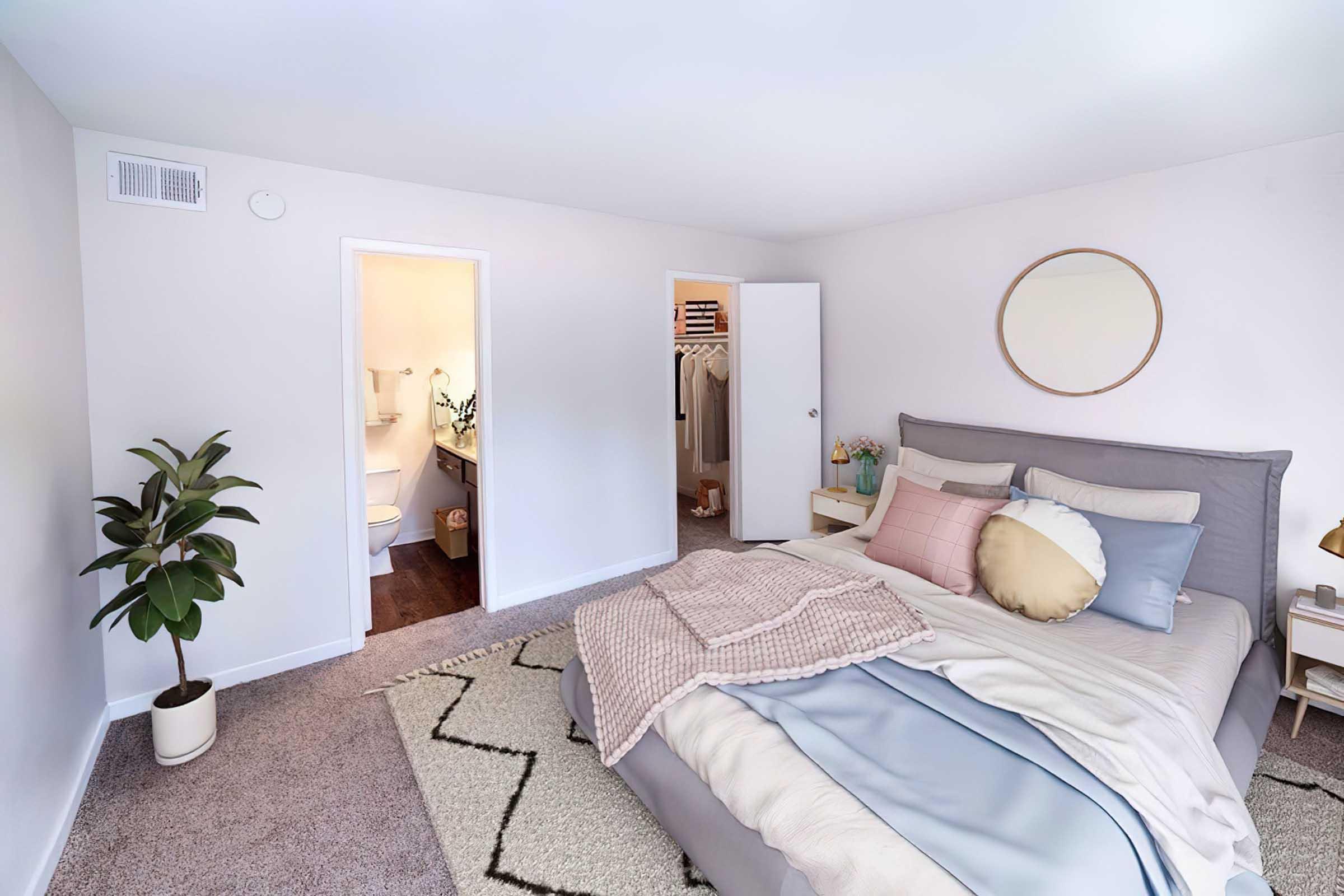 A cozy, well-lit bedroom featuring a gray bed with pastel-colored pillows and a soft blanket. A round mirror hangs on the wall, and a potted plant adds greenery. A door leads to a bathroom, and another door opens to a closet area, enhancing the room's inviting atmosphere.