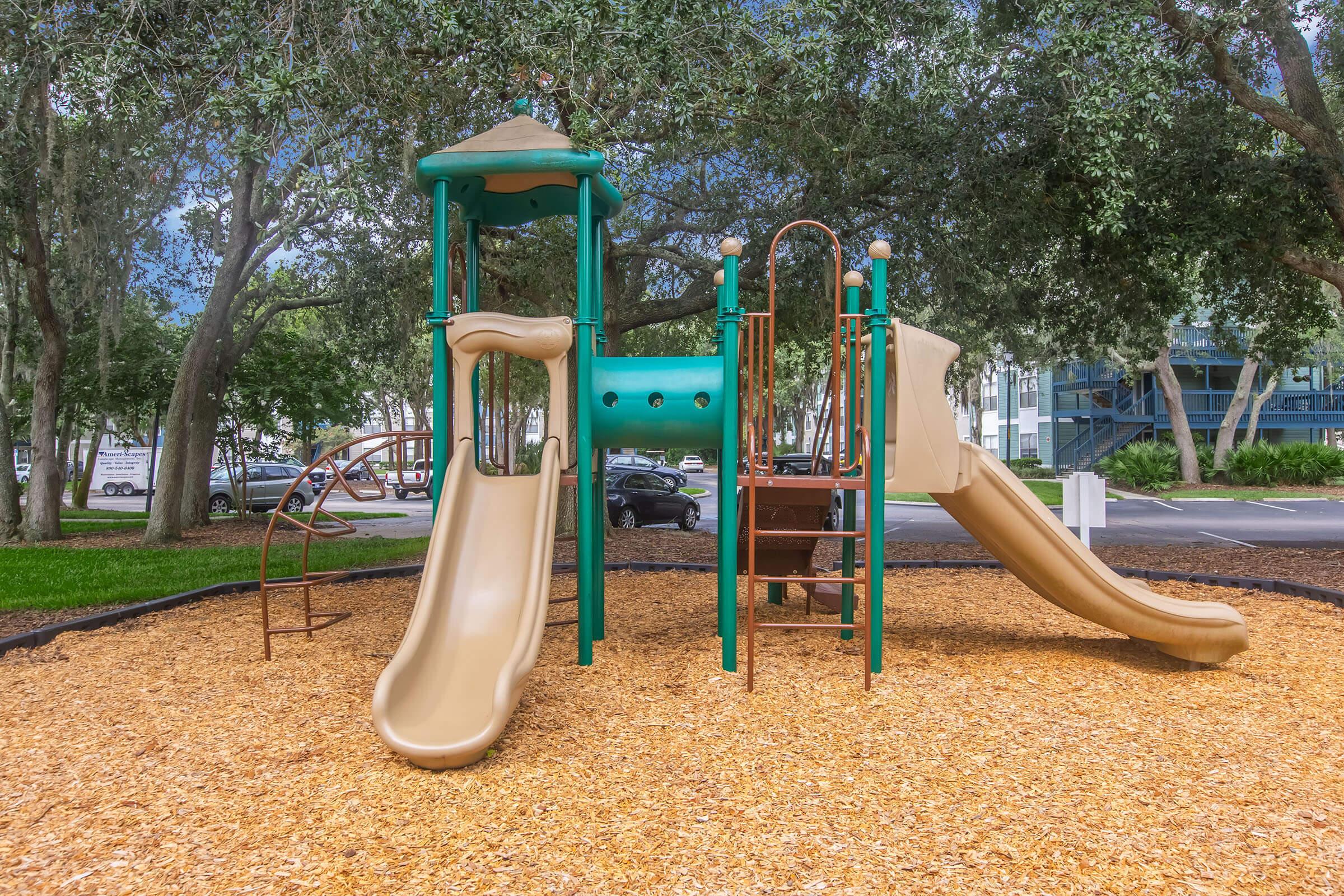 A colorful playground structure featuring two slides, a climbing area, and a small roof. The playground is situated on a bed of tan wood chips, surrounded by trees, with a parking lot visible in the background.
