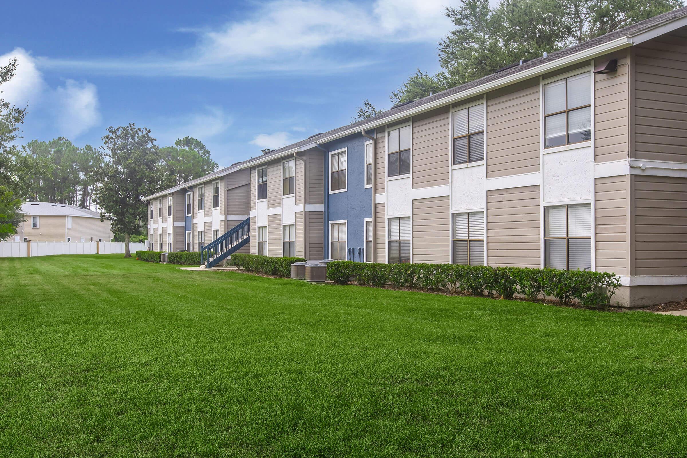 A side view of a two-story apartment building featuring a mix of beige and blue exterior panels. The well-maintained lawn is lush and green, with neatly trimmed bushes along the foundation. Behind the building, additional structures and trees are visible against a clear blue sky.