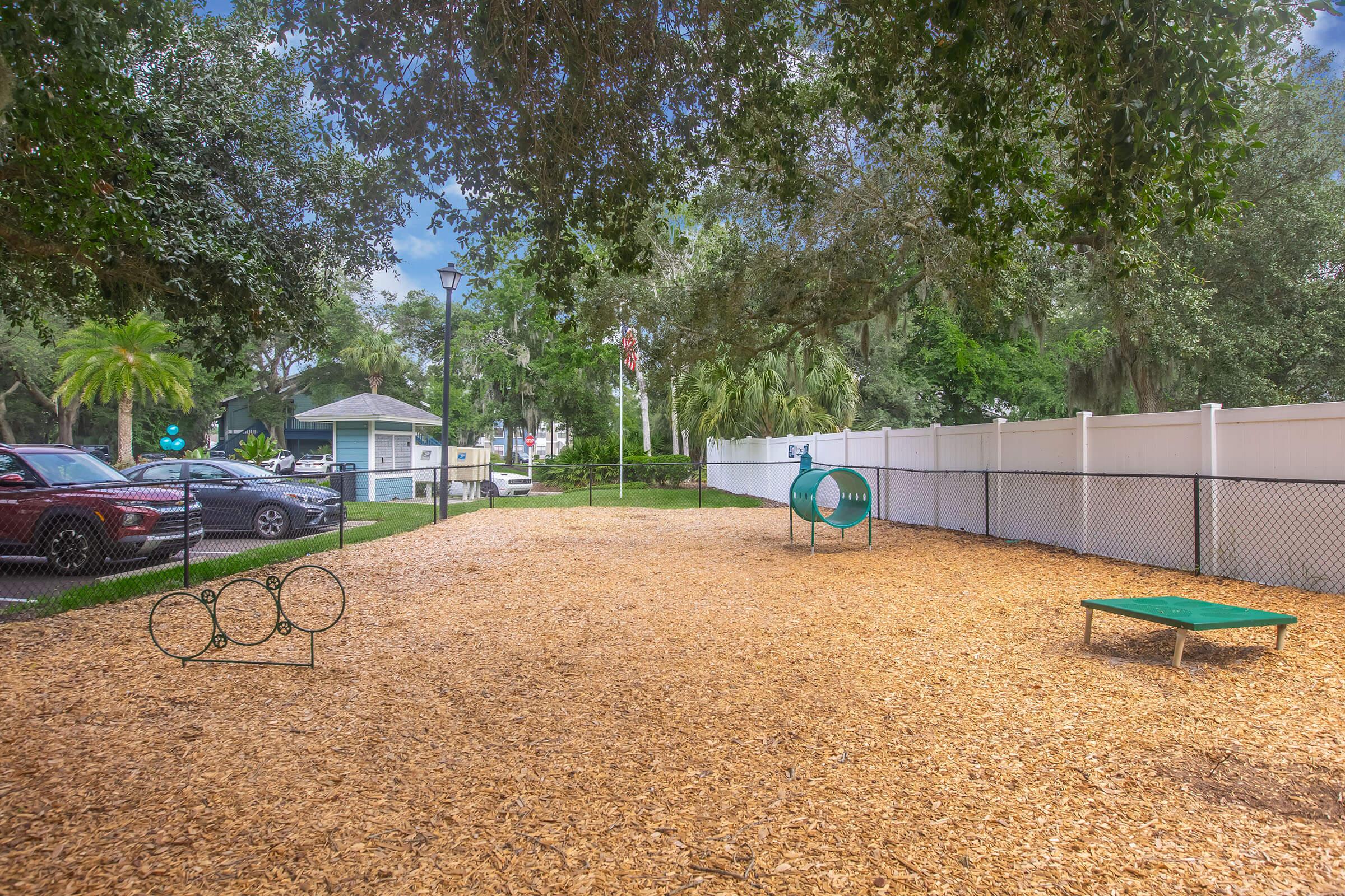 A dog park with wood chip flooring, featuring a green play structure and a small agility ramp. Surrounded by a white fence and trees, the park includes benches and is near buildings and parked cars. A calm atmosphere with a partly cloudy sky.