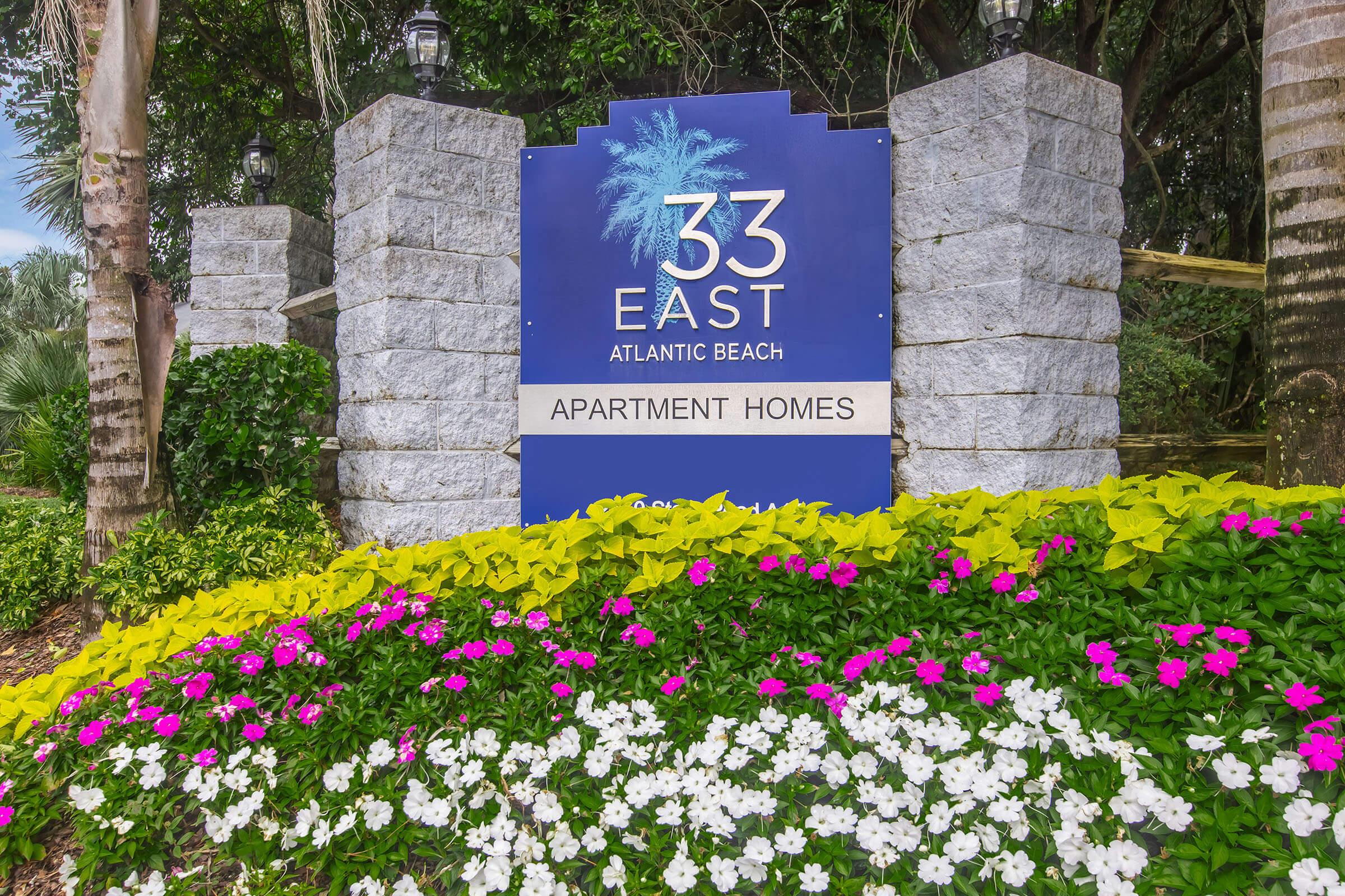 Sign for "33 East Apartment Homes" at Atlantic Beach, featuring a blue background with white lettering and a palm tree graphic. The sign is flanked by stone pillars and surrounded by colorful flower beds with pink and white blooms, creating a welcoming atmosphere.