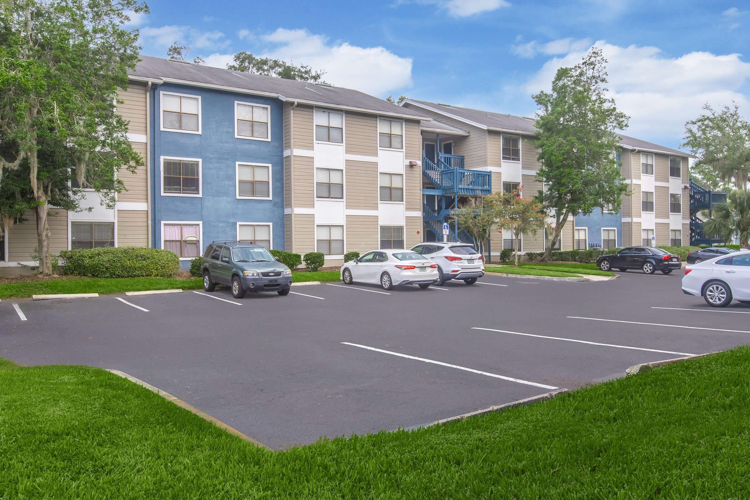 A view of a multi-story apartment complex featuring a mix of beige and blue exterior. The foreground shows a parking lot with various vehicles, surrounded by greenery and well-maintained landscaping. The sky is clear with a few clouds, indicating a pleasant day.