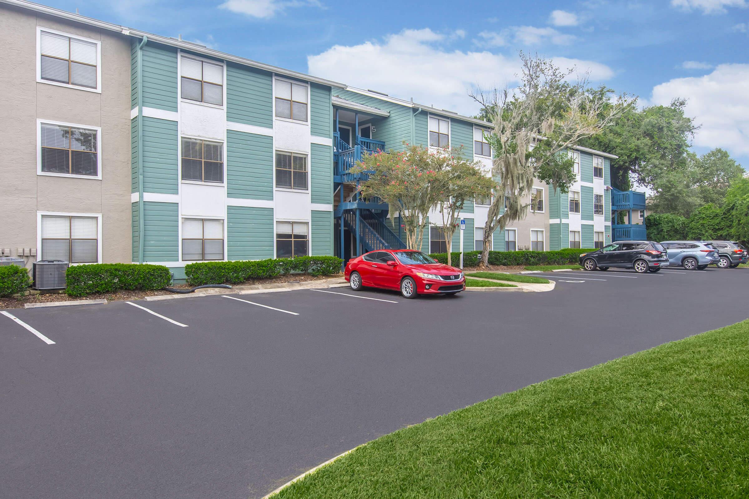 A residential apartment complex featuring a two-story building with light blue and white siding. A red car is parked in the foreground, alongside several other vehicles. Lush green grass and shrubs line the parking lot, while trees are visible nearby under a clear blue sky.