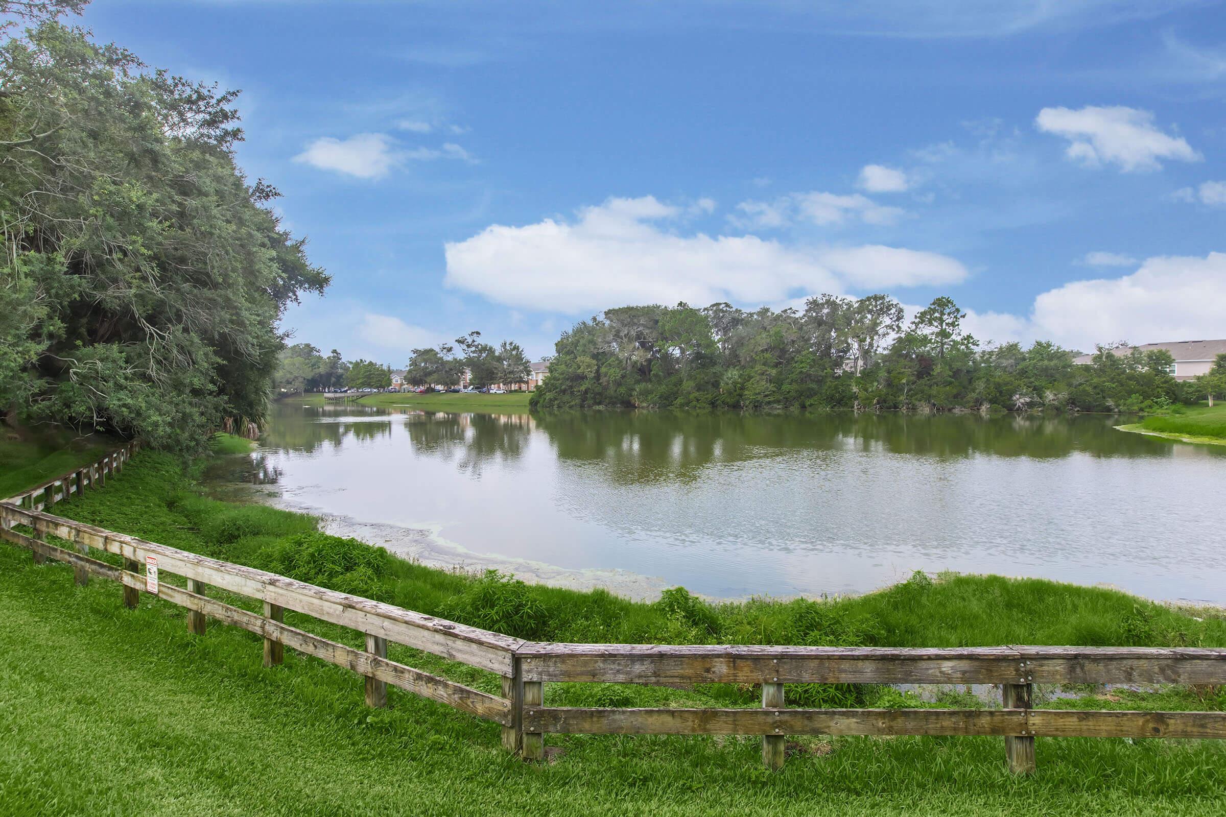 A serene landscape featuring a calm lake surrounded by lush greenery and trees. There is a wooden fence along the bank of the lake, and the sky above is bright with scattered clouds. The scene reflects a tranquil outdoor setting, perfect for relaxation and nature appreciation.