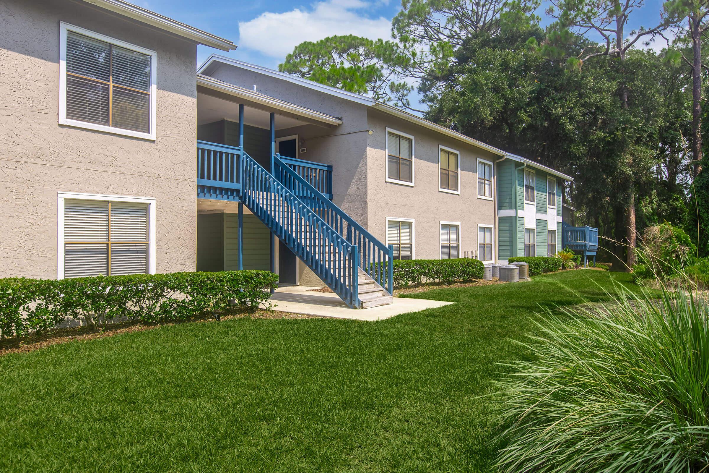 Exterior view of a two-story apartment building with light gray walls and blue railings. There are well-maintained grassy areas and shrubbery in the foreground. The building features multiple windows, a staircase leading to the second floor, and is surrounded by trees in a sunny environment.