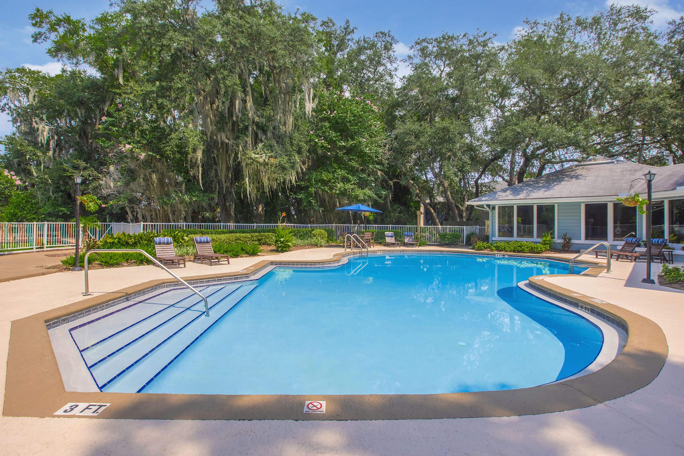 A serene outdoor swimming pool surrounded by lush greenery. The pool features a curved shape with steps leading into the water, and several lounge chairs are positioned nearby. A small building with a shaded area can be seen in the background, creating a relaxing atmosphere for leisure and recreation.