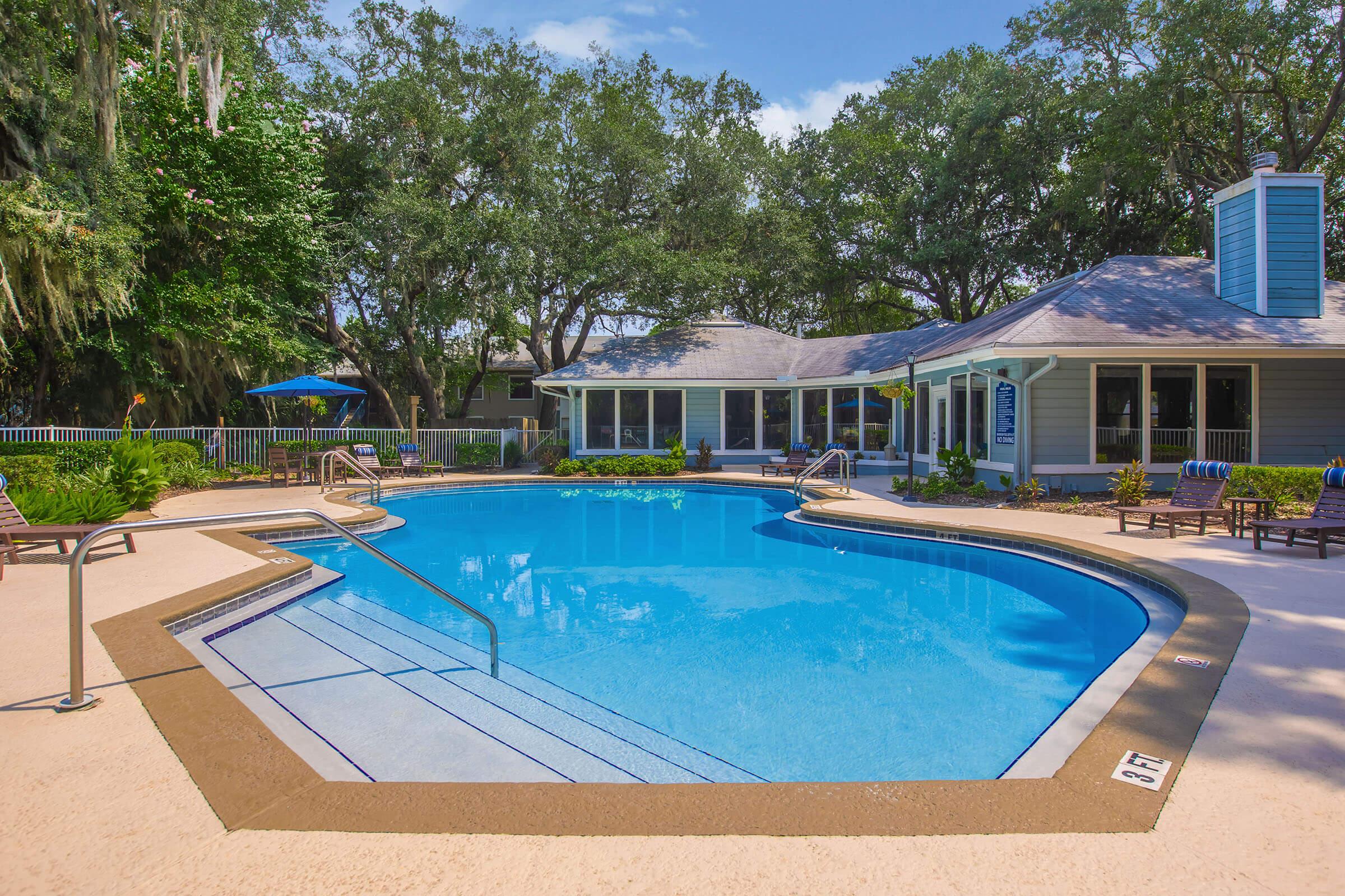 A clear blue swimming pool surrounded by a concrete deck, lined with lounge chairs and an umbrella. Lush green trees provide a natural backdrop, and a residential building with large windows and a chimney is visible in the background. The scene depicts a tranquil outdoor setting perfect for relaxation.