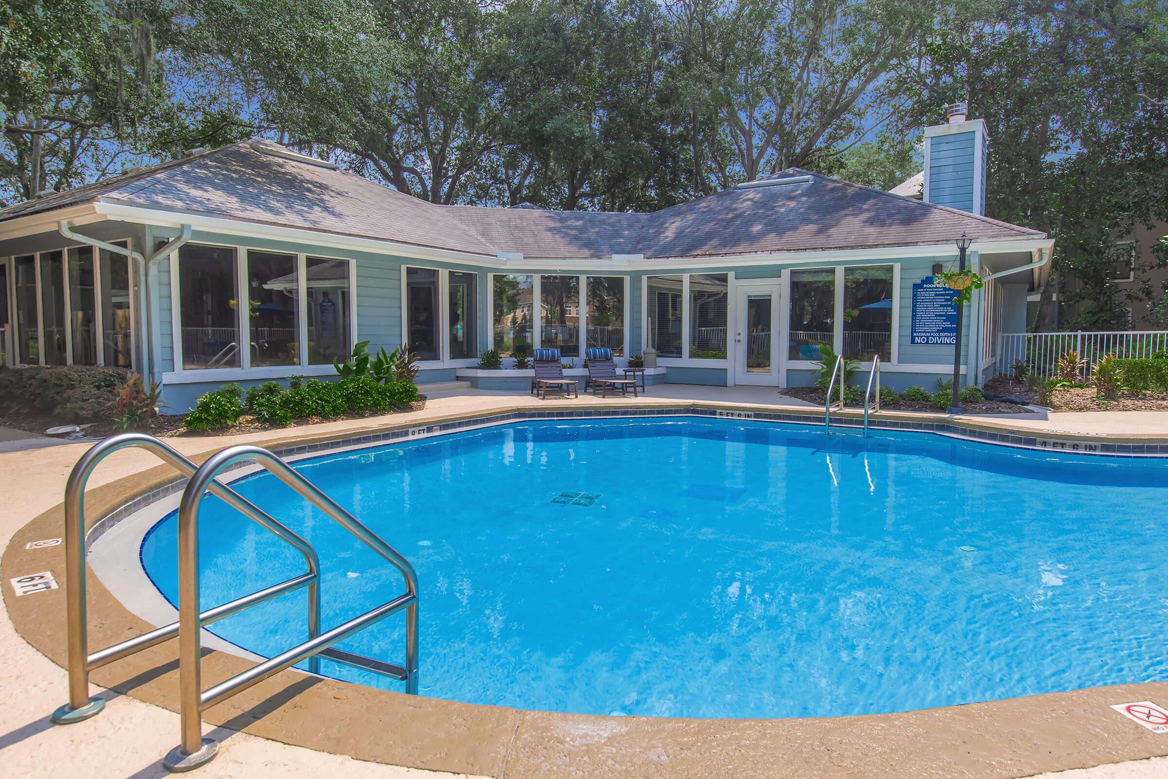 A residential pool area featuring a large, blue swimming pool with a ladder, surrounded by a concrete deck. In the background, there is a modern house with large windows and a patio, framed by lush greenery and trees. The setting is well-lit, suggesting a sunny day.