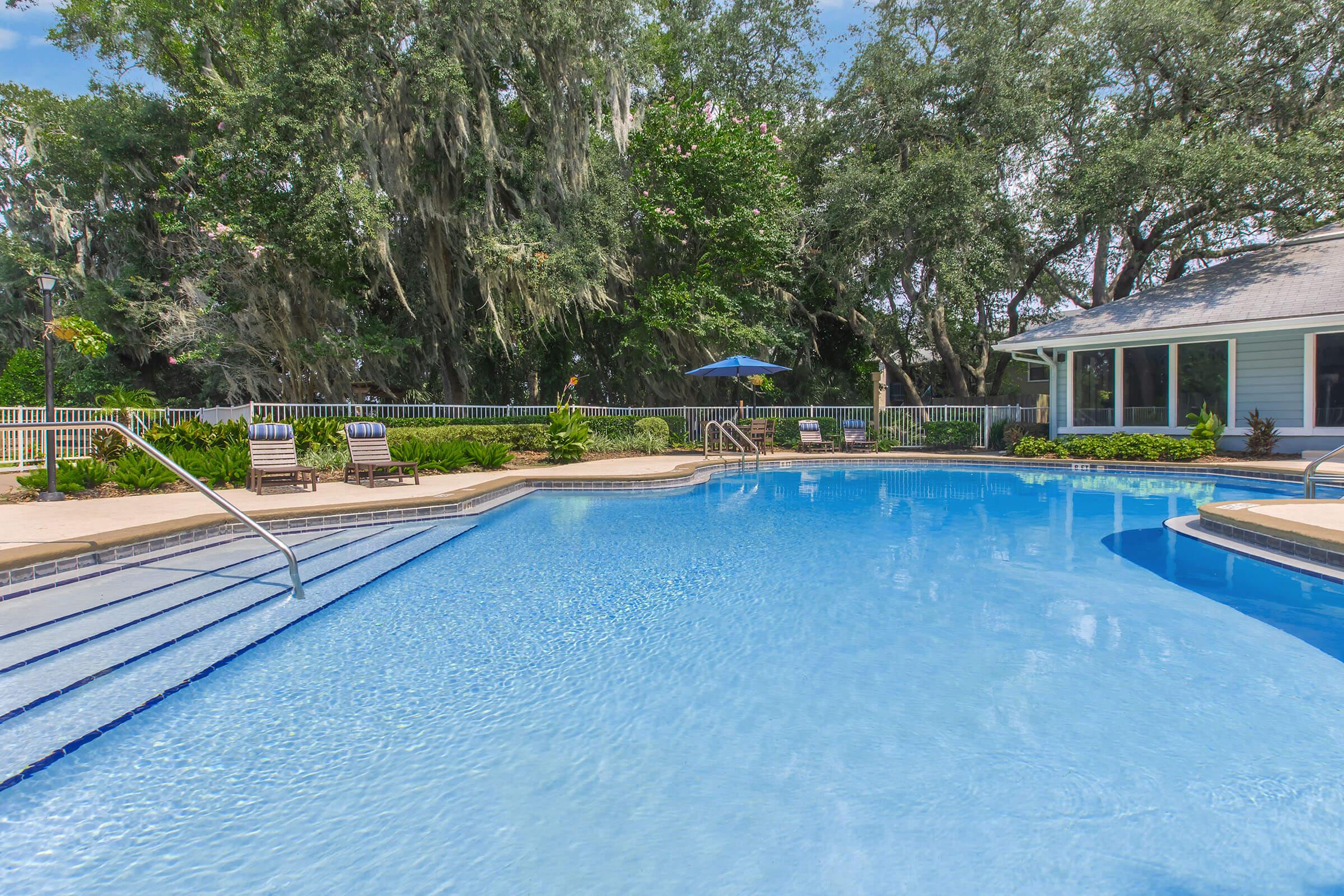 A tranquil swimming pool surrounded by lush greenery and trees, with comfortable lounge chairs nearby. The pool features clear blue water and steps leading in, while a small building with large windows and a blue umbrella adds to the relaxing atmosphere.