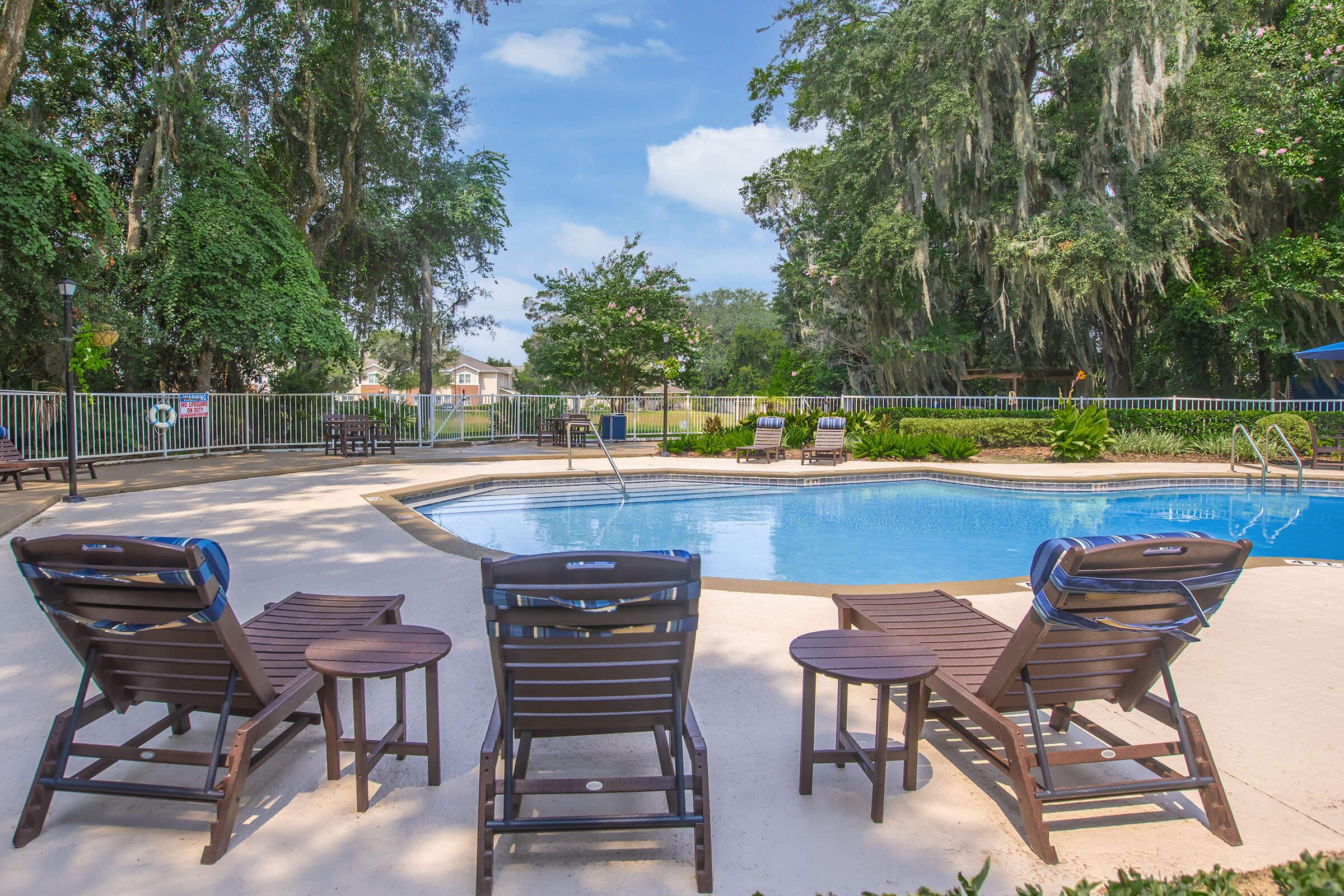A serene pool area surrounded by lush greenery. Four wooden lounge chairs are positioned near the edge of the pool, which reflects the blue sky above. Tall trees and plants provide a natural backdrop, creating a relaxing environment for sunbathing and leisure.