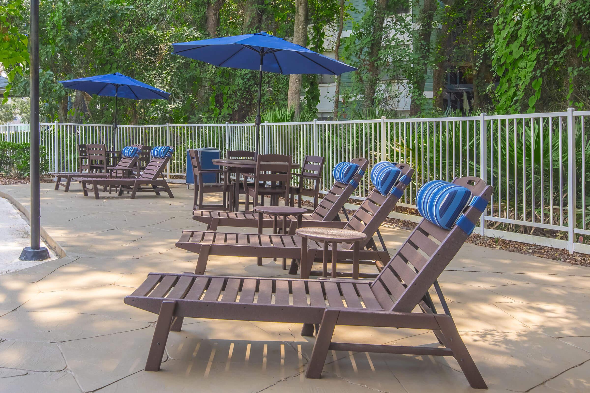 Outdoor seating area featuring wooden lounge chairs with blue and white striped cushions, surrounded by lush greenery. There are several blue umbrellas providing shade over the chairs and a small table nearby, creating a relaxing atmosphere by a poolside.