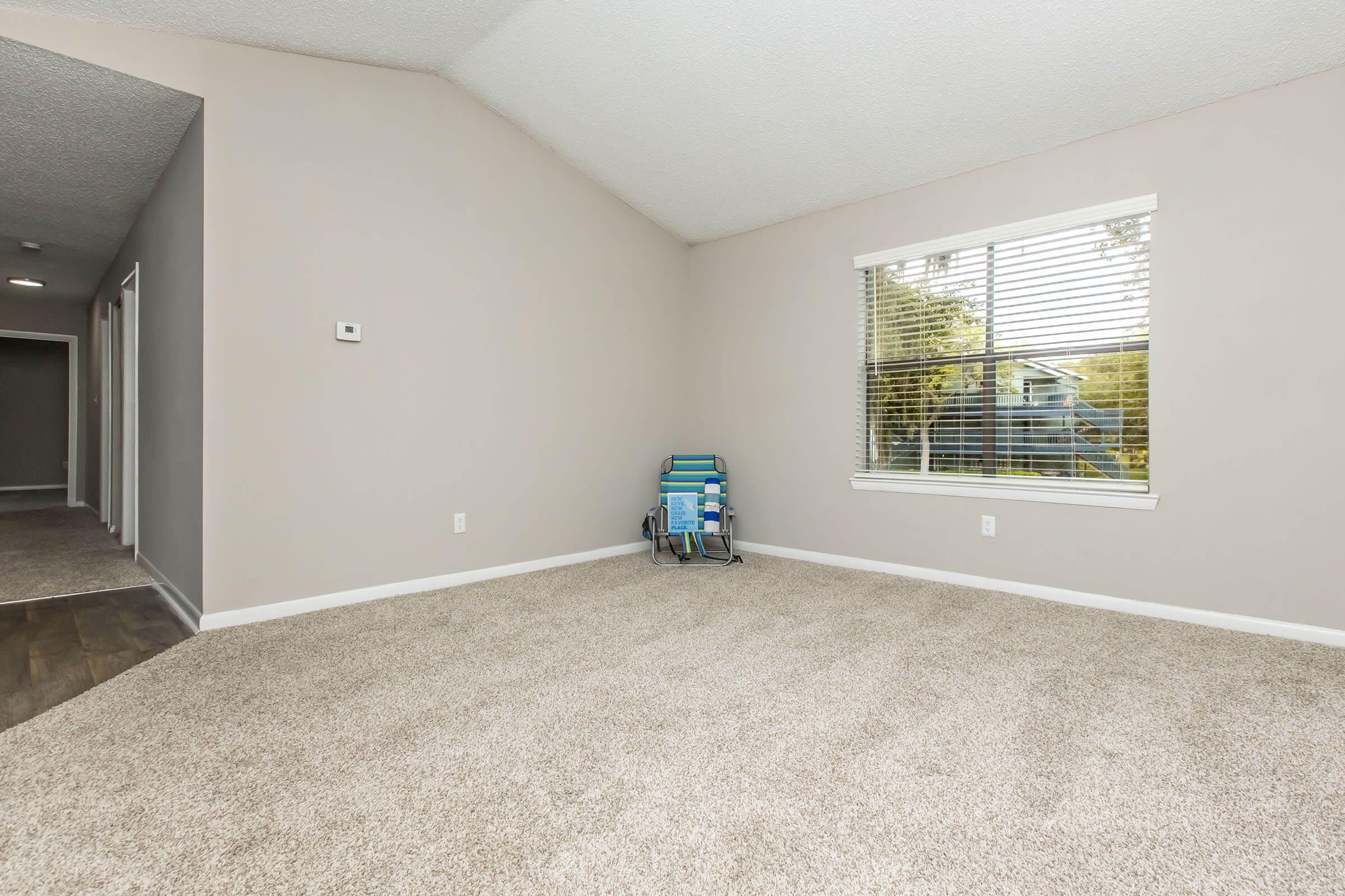 A spacious, empty room with light-colored walls and a carpeted floor. There is a window with blinds on the right side, allowing natural light to enter. In the corner, there is a blue and green folding chair. Opposite the window, a hallway with doors suggests access to other rooms.