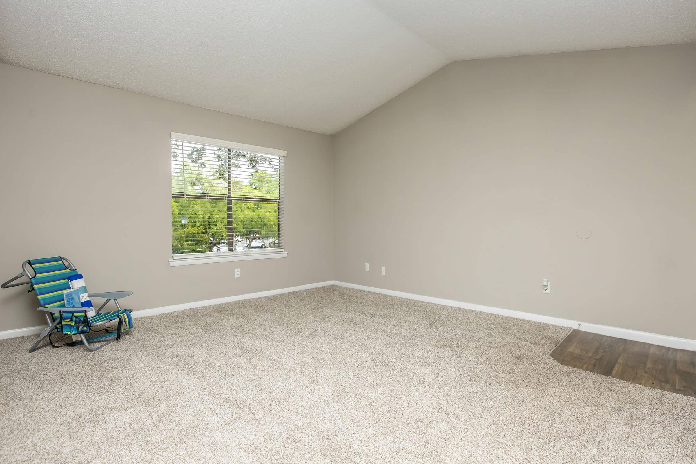 A spacious, empty room with light beige carpet and gray walls. A window with white blinds allows natural light in, overlooking greenery outside. In one corner, there is a blue and green folding chair. The room features a high, sloped ceiling and has minimal decor, creating a clean, open atmosphere.