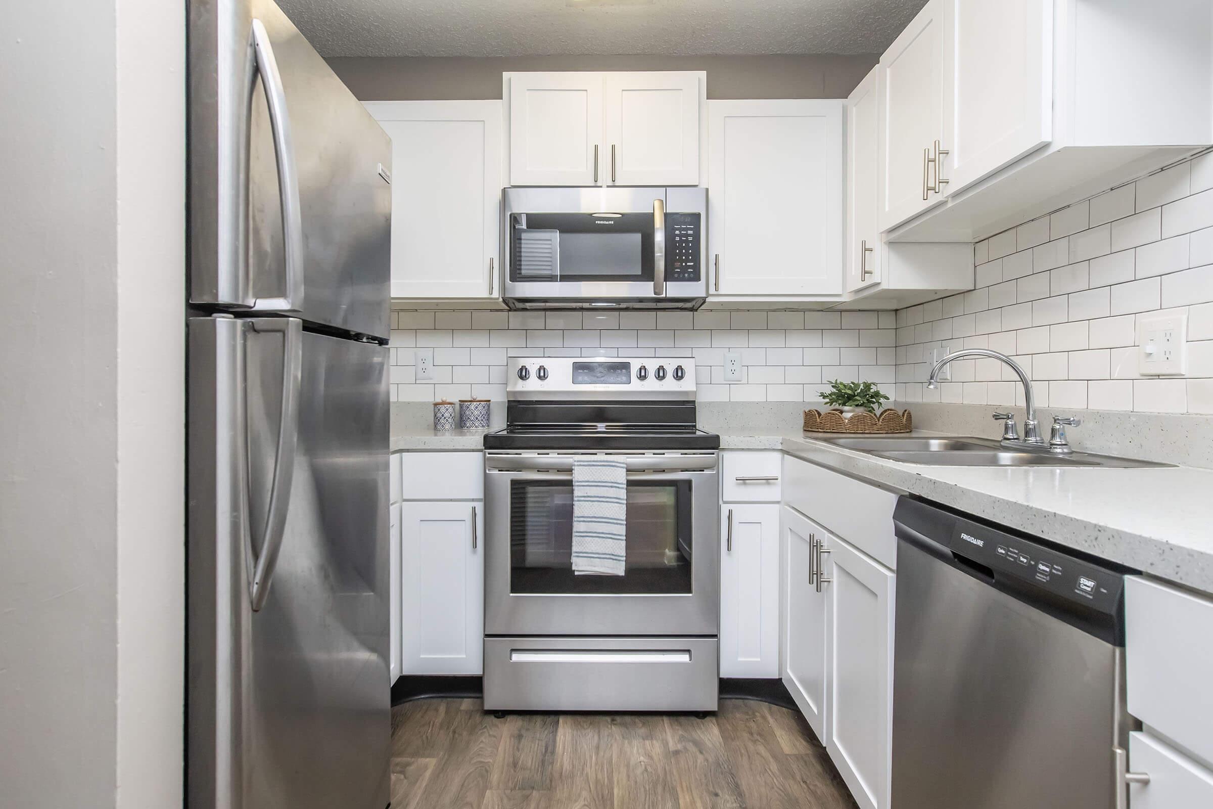 Modern kitchen featuring stainless steel appliances, including a refrigerator, microwave, and oven. Light-colored cabinets and a white subway tile backsplash complement the countertops. A sink is positioned next to a dishwasher, with a small plant display on the counter. Hardwood-style flooring adds warmth to the space.