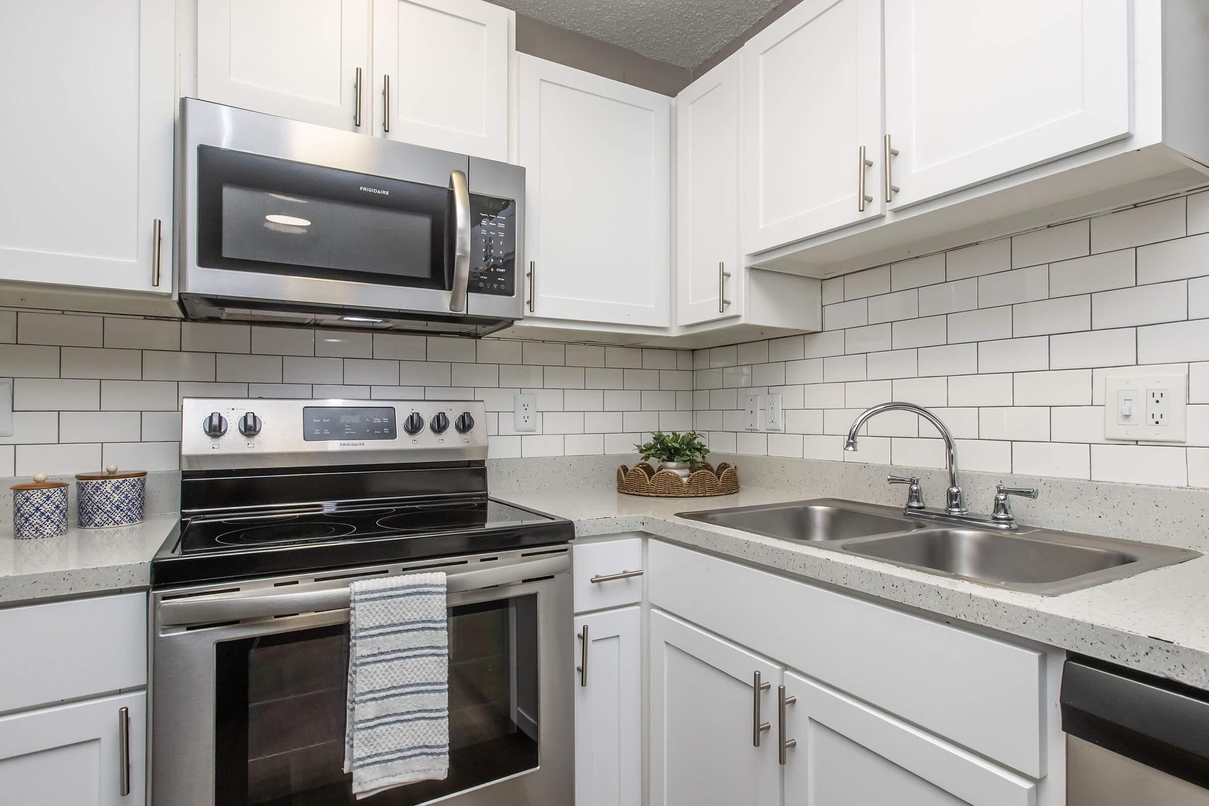 Modern kitchen with white cabinetry, stainless steel appliances, including a microwave and an oven. Features a gray countertop and a double sink with a faucet. Backsplash consists of white subway tiles. Three decorative containers are placed on the counter, along with a small potted plant.
