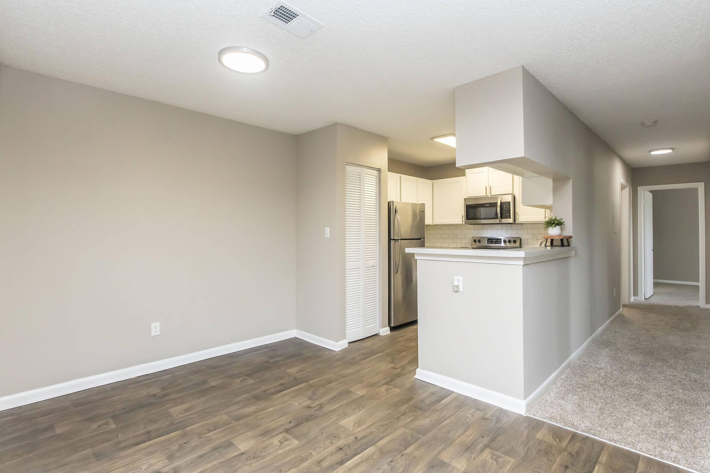 A spacious and modern living area with light gray walls and hardwood flooring. The kitchen features stainless steel appliances, including a microwave and refrigerator, with a breakfast bar. The adjoining space is carpeted and leads to other rooms, contributing to an open and inviting atmosphere.