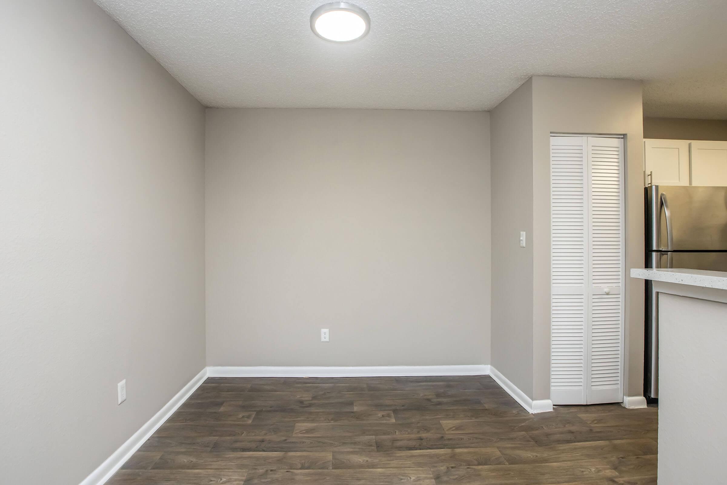 An empty room with light gray walls and a medium wood-like floor. A round ceiling light fixture illuminates the space. There is a white closet door on the right side, and a kitchen area is partially visible in the background. The room appears clean and freshly painted, ideal for decoration or furniture placement.