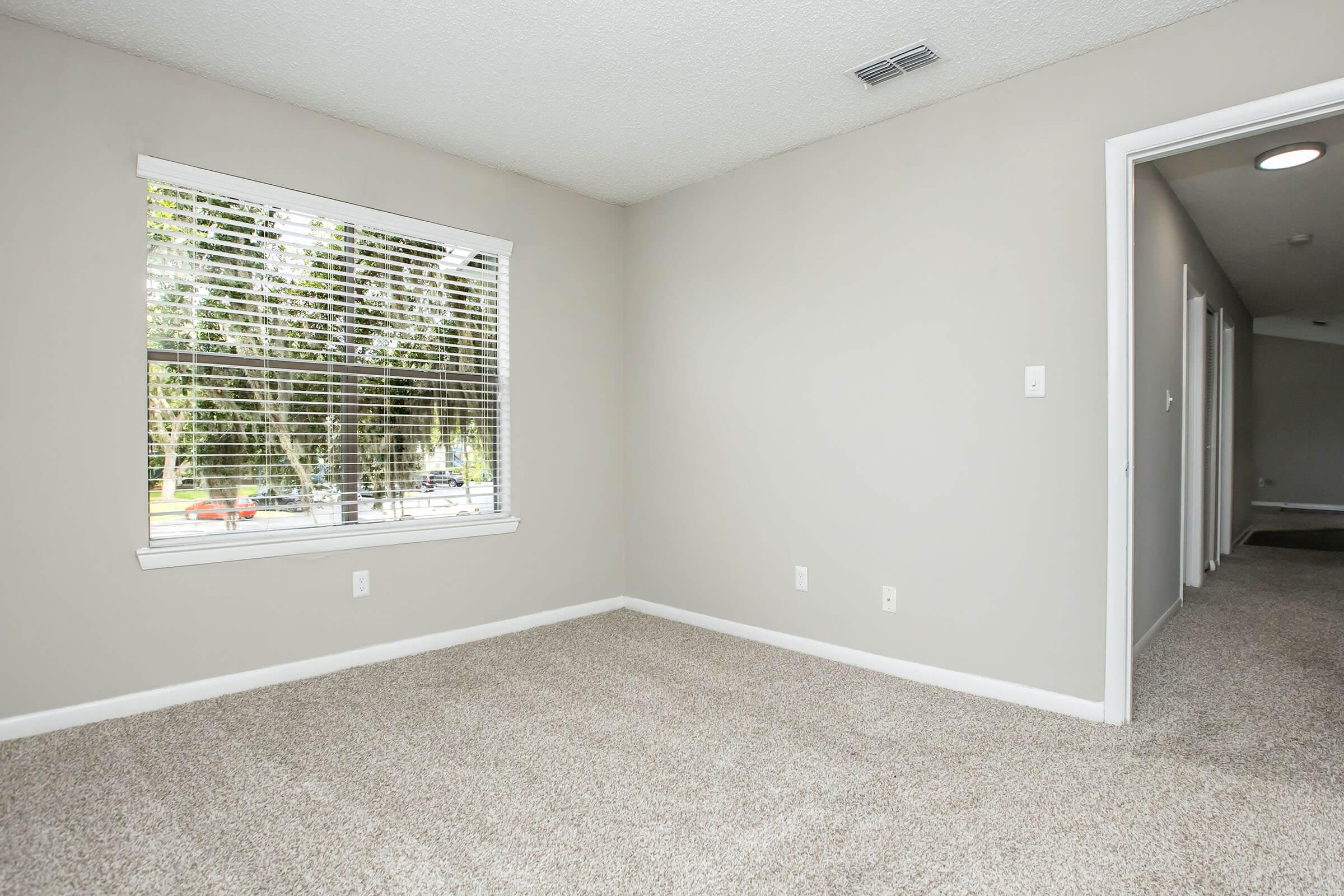 A bright, empty room with light gray walls and carpet. A window with blinds allows natural light in, revealing a view of trees and a street outside. On one side, a hallway leads to other areas of the house, and the space appears clean and well-maintained.
