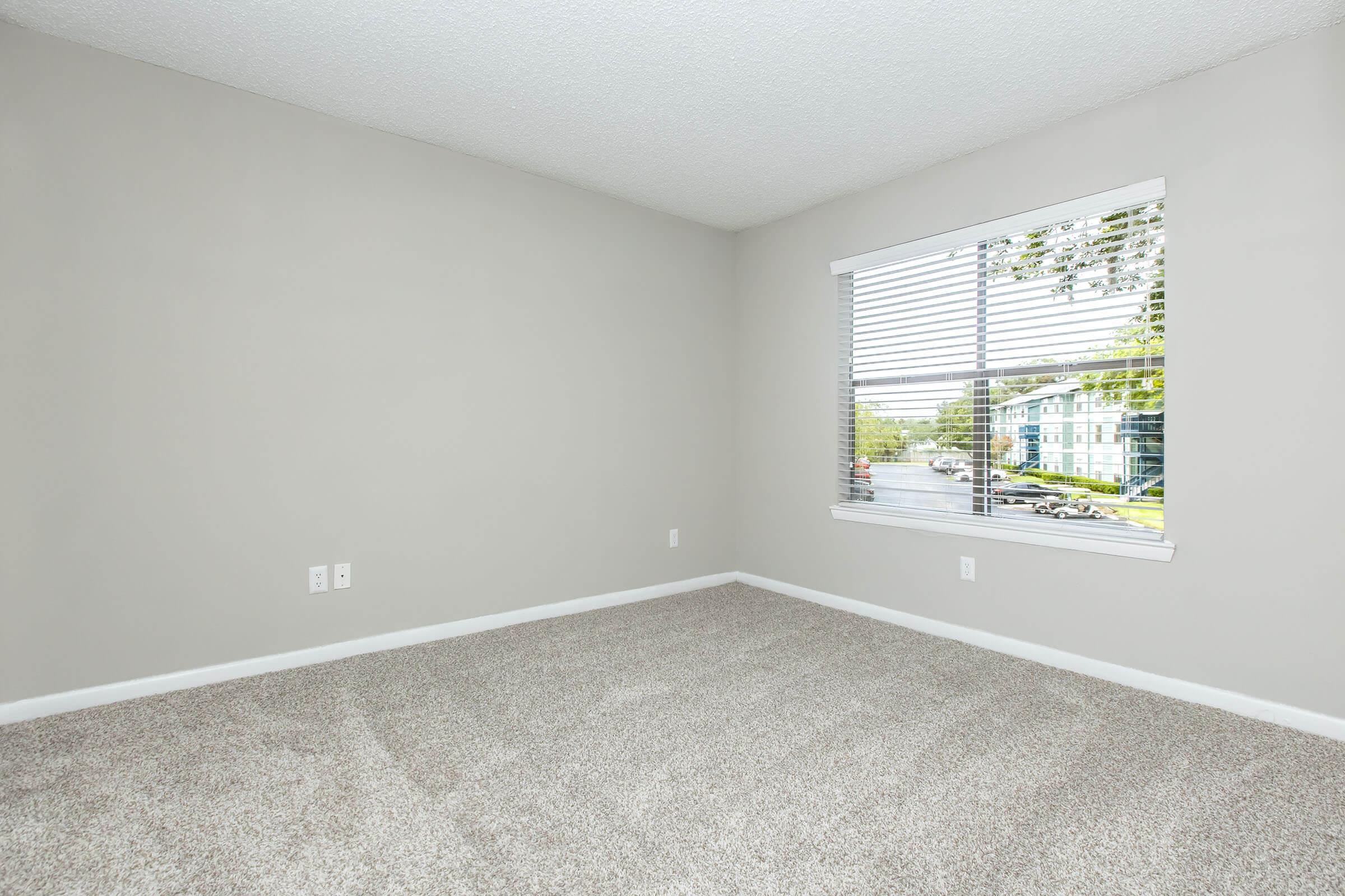 A vacant room with light gray walls and a carpeted floor. There is a window with white blinds, allowing natural light to enter the space. Outside the window, a view of a street and buildings is visible. The room appears clean and ready for moving in.