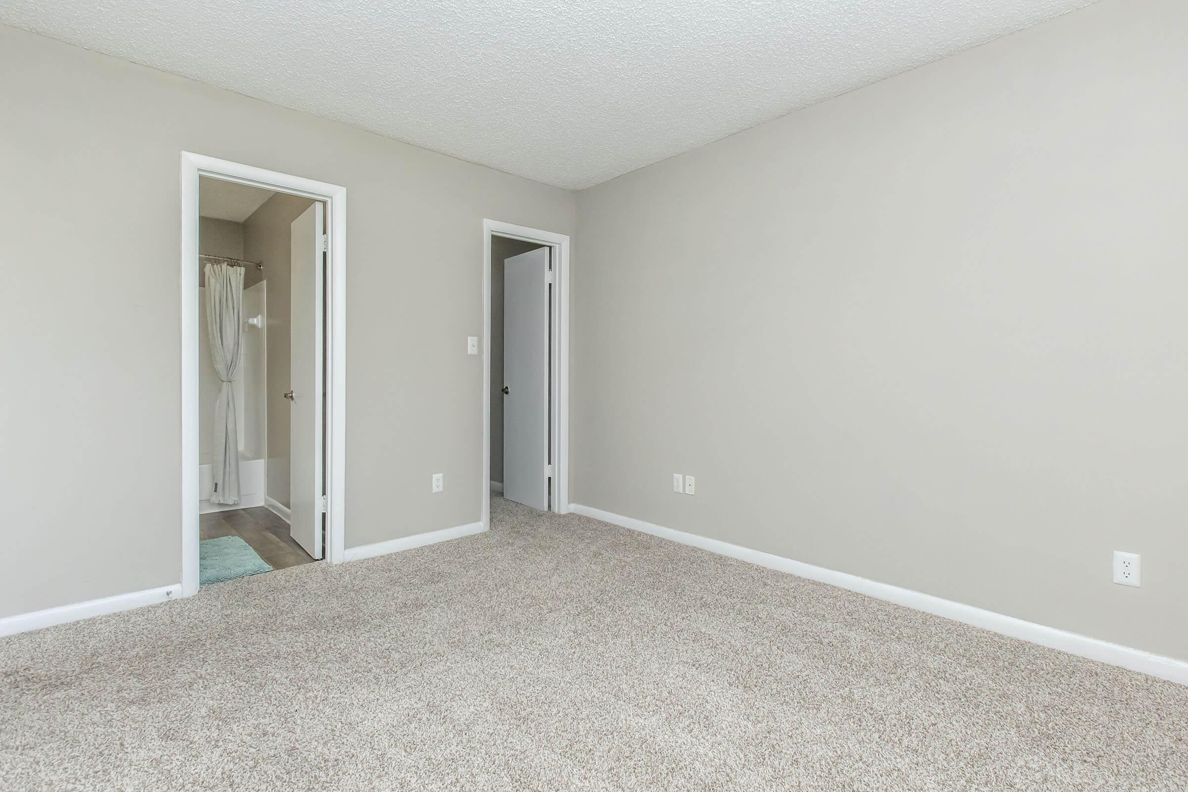 A vacant, well-lit room with beige carpeting and light gray walls. There are two doors—one leading to a bathroom and the other to a hallway. The room has no furniture, emphasizing the spaciousness and neutral decor.